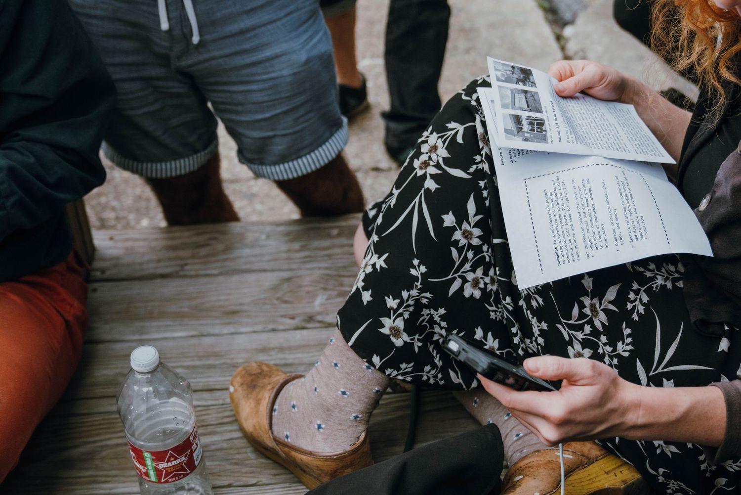 Person in floral skirt reading a newsletter while sitting on wooden deck with water bottle nearby.