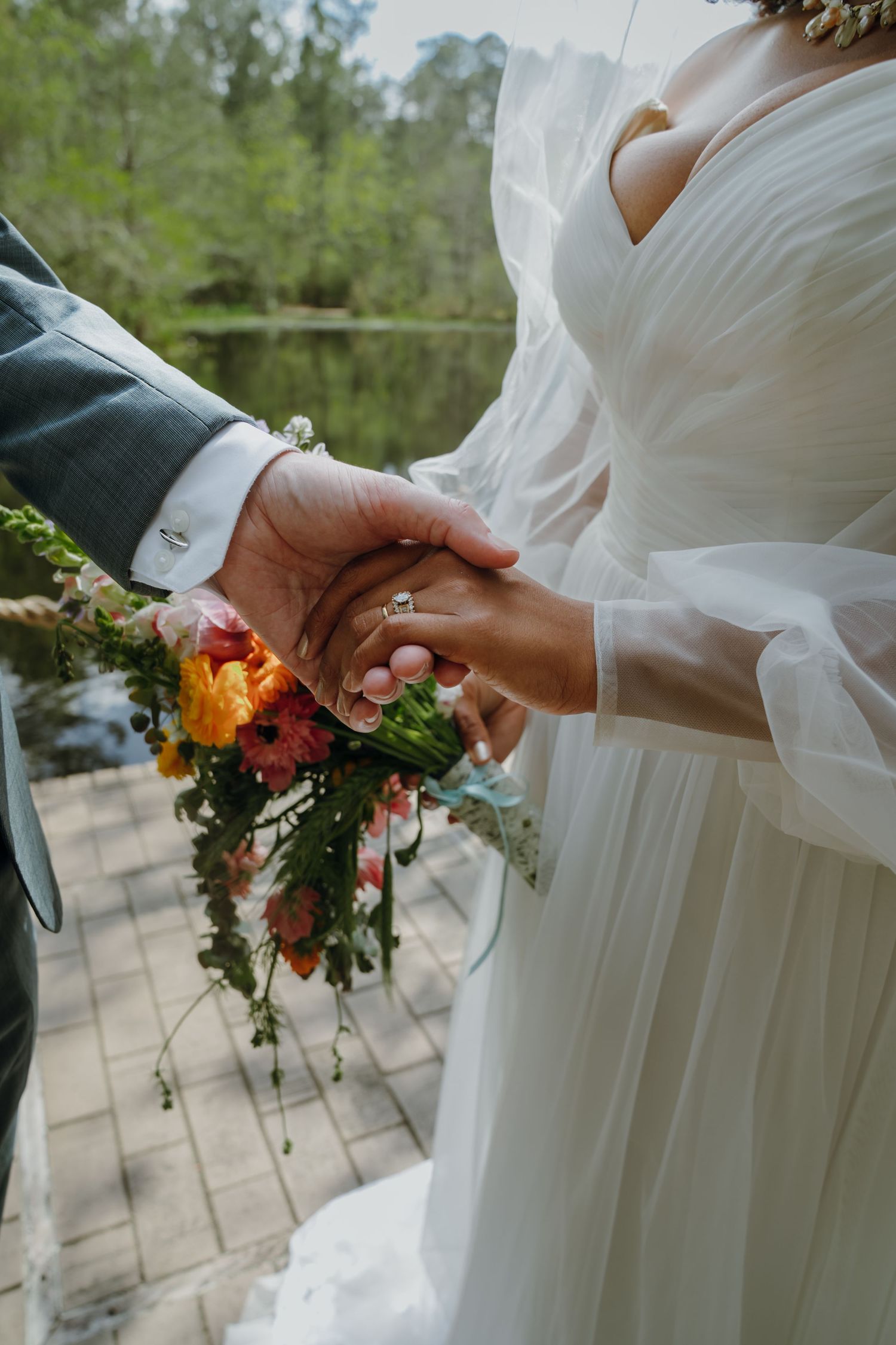 Close-up of bride and groom holding hands with colorful bridal bouquet by lakeside.