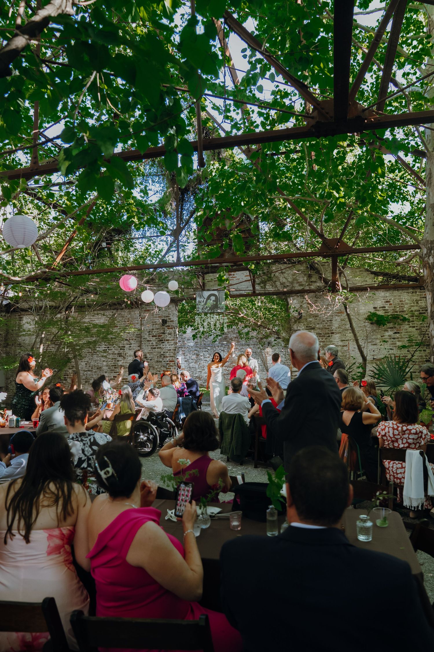 Outdoor wedding reception under lush green tree canopy with guests seated at tables and couple celebrating near stone wall.