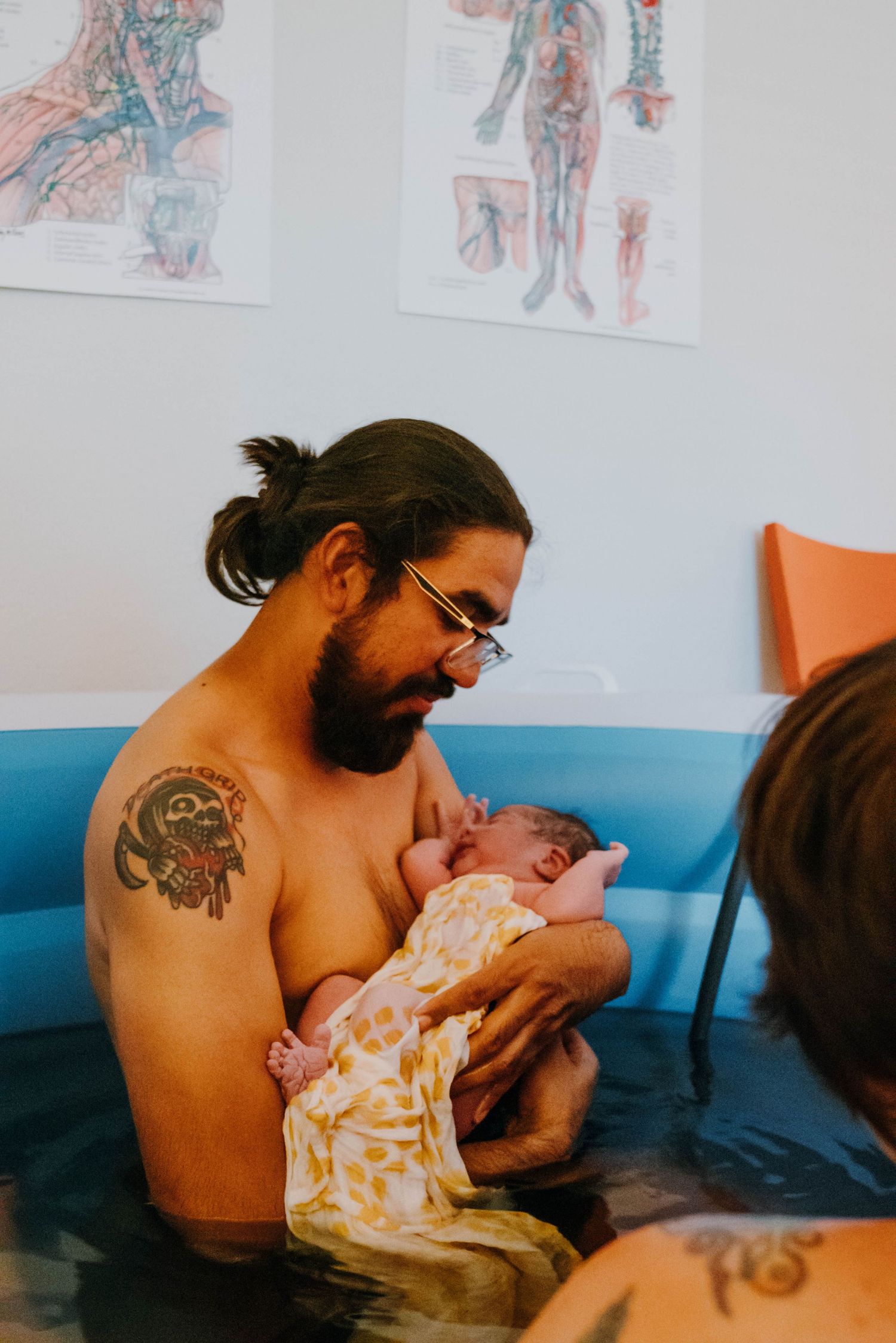 Tattooed father cradles newborn baby in arms while sitting in a home water birth pool.