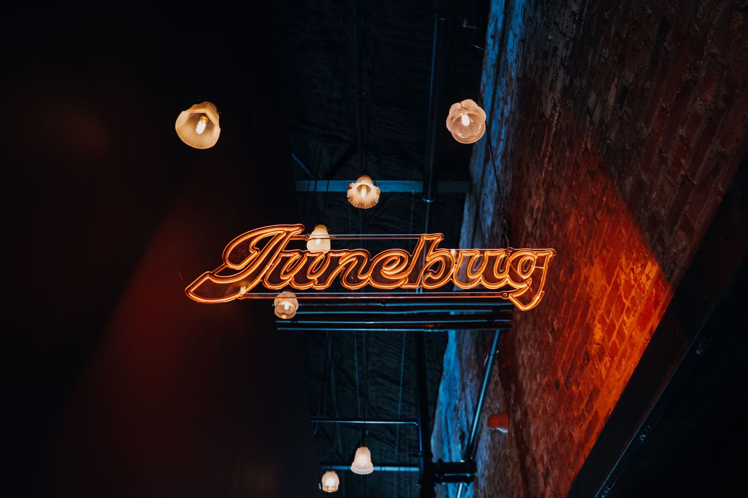 Glowing neon Junebug sign hanging from a brick ceiling with vintage Edison bulb pendants.