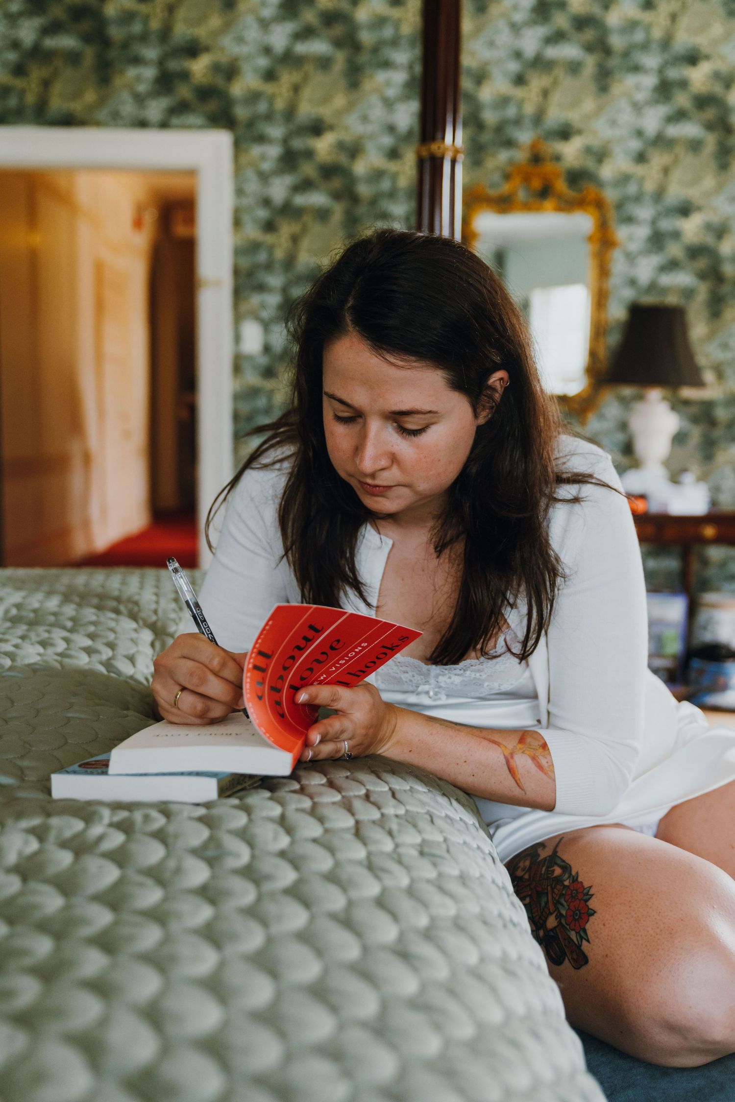 Bride in white robe writing in a red book while sitting on a bed in an elegant floral wallpapered room. New Orleans, Louisiana, Mississippi