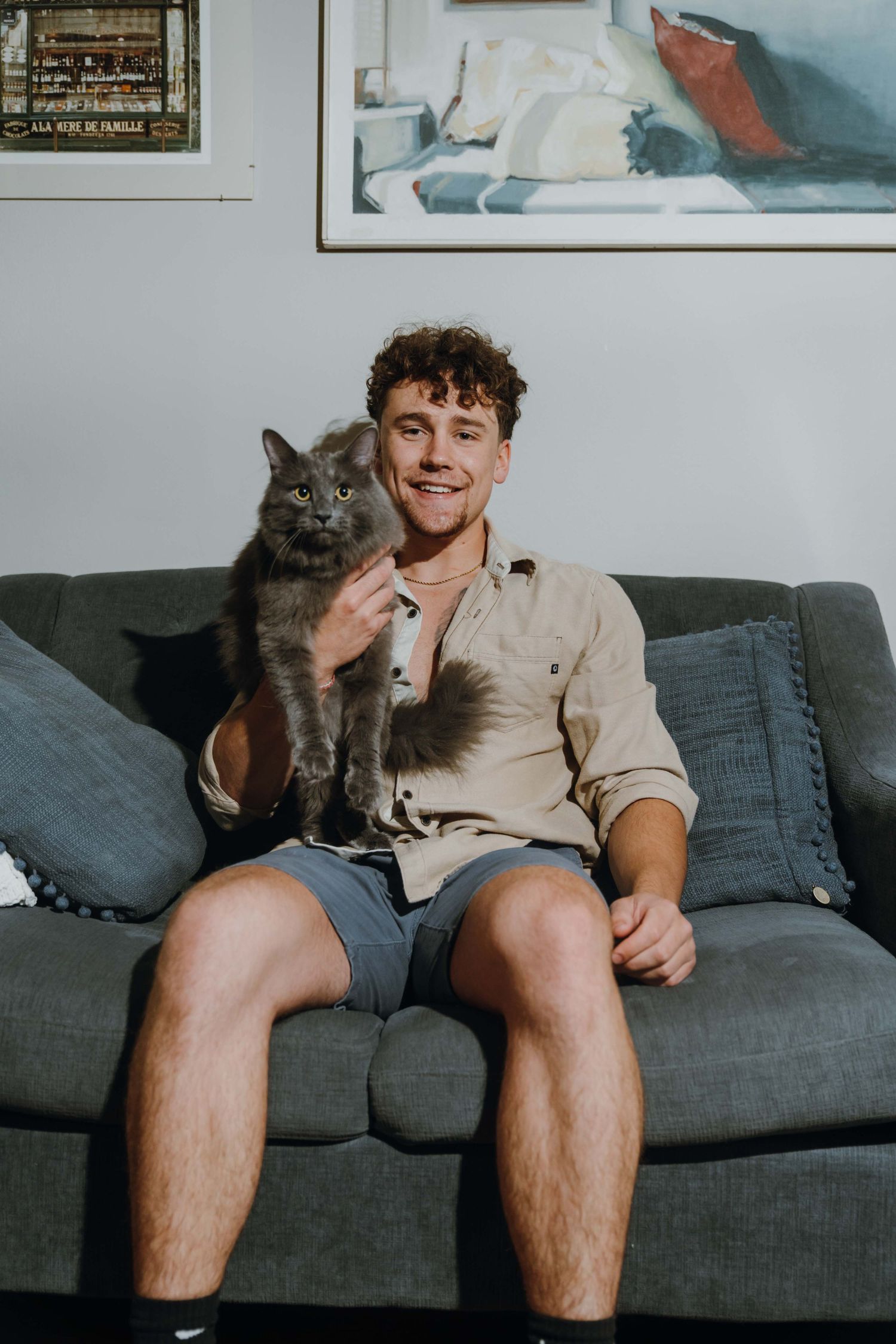 Young man in beige shirt and shorts sitting on gray sofa holding a fluffy gray cat, with artwork on wall behind.