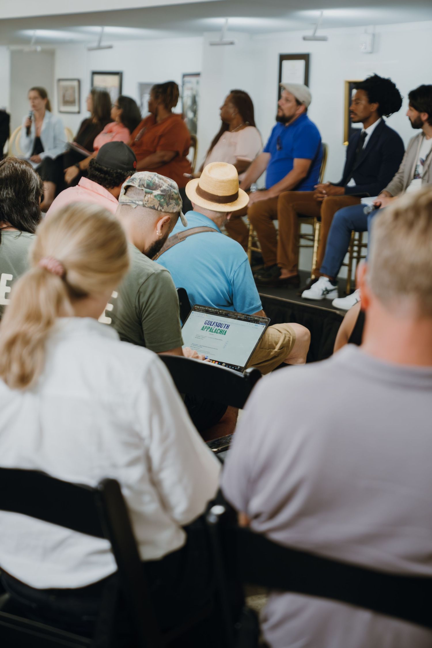 Diverse group of people seated in rows at a community panel discussion event in a bright white room.
