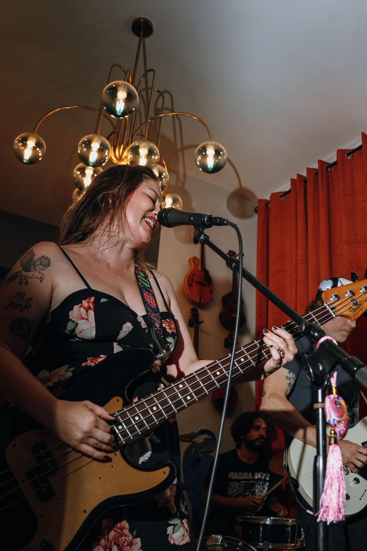 Woman with tattoos playing bass guitar and singing at a live indoor concert under a gold chandelier.