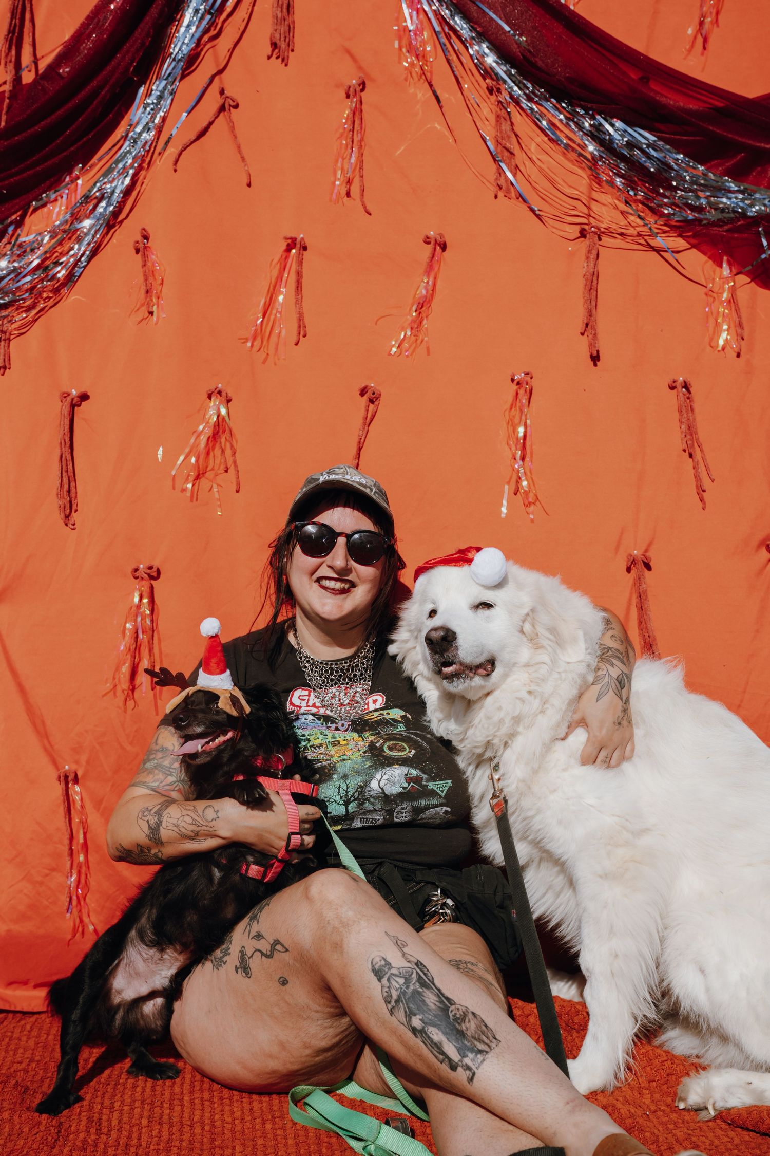 Tattooed woman in sunglasses sits with two dogs wearing Santa hats against festive orange backdrop.