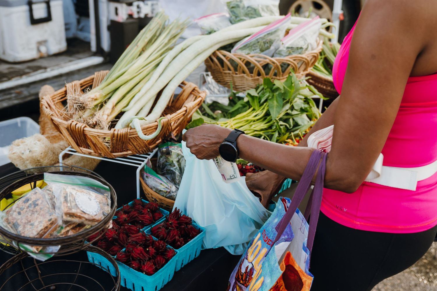 Woman in pink top shopping for fresh vegetables and berries at an outdoor farmers market stand.
