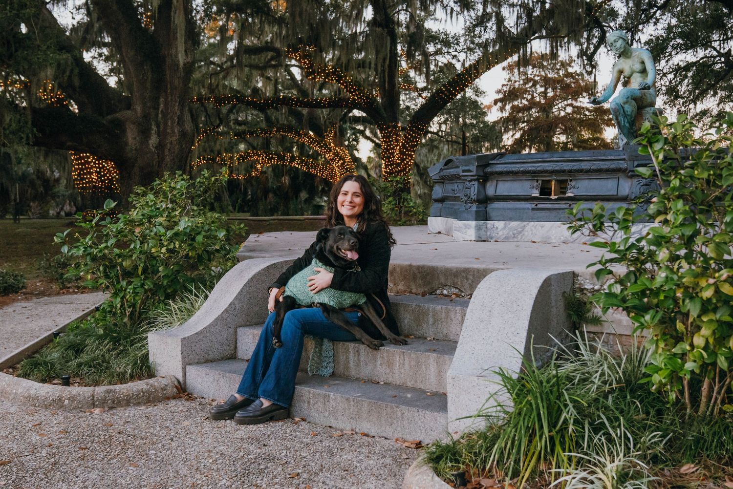 Woman sitting on stone steps in a lush garden park, holding a small dog near a statue.