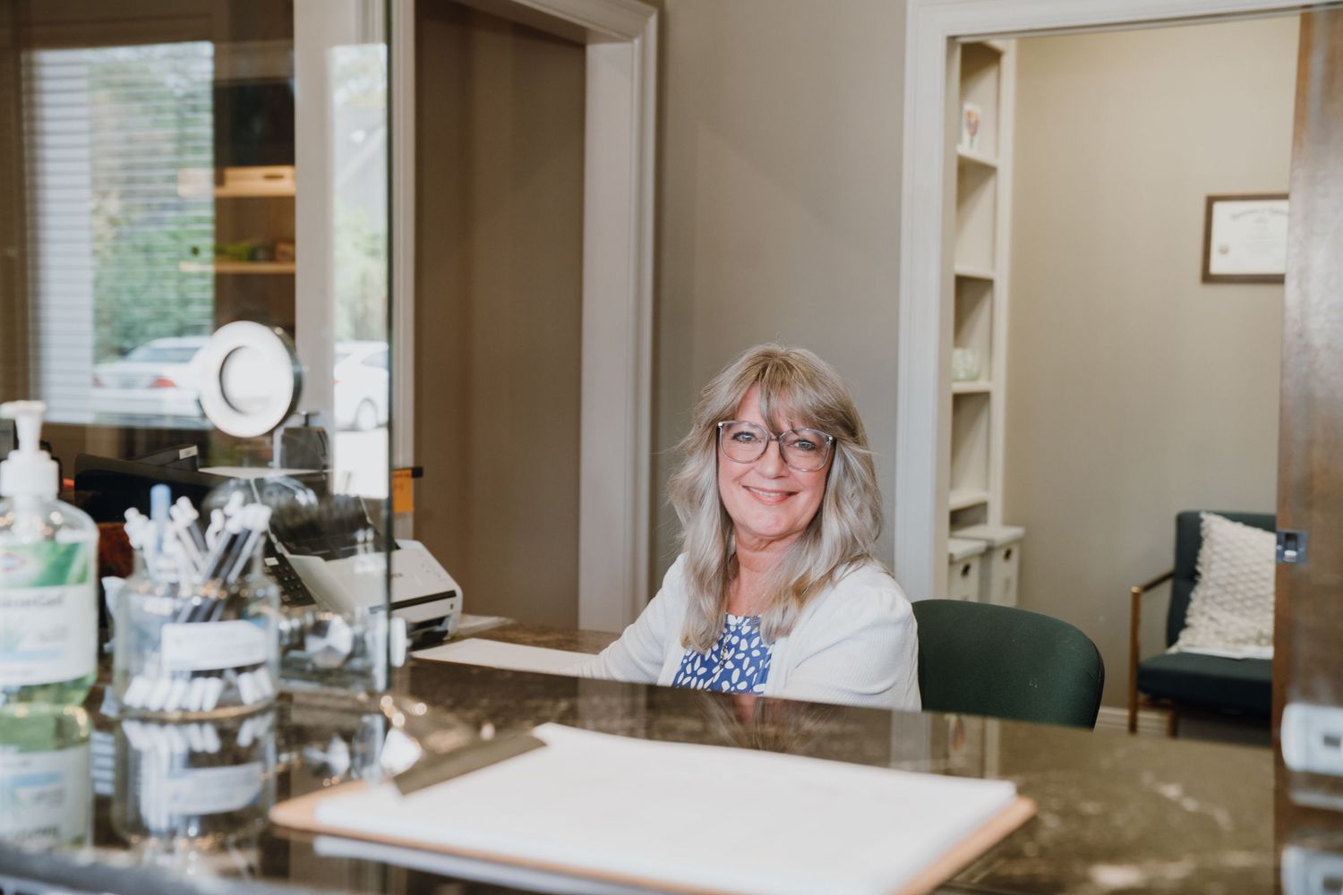 A friendly receptionist with glasses smiles warmly at the front desk of a dental office.