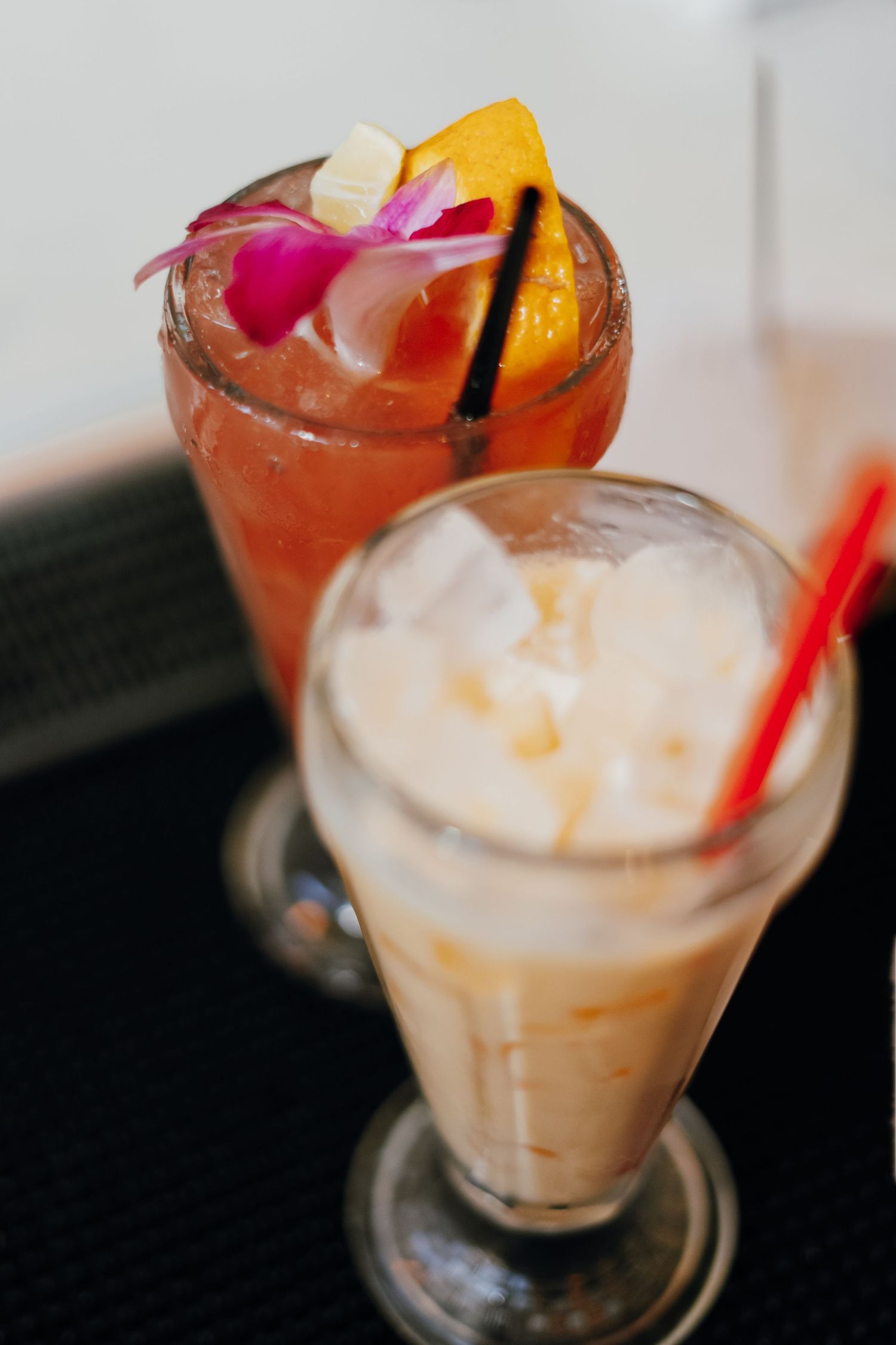 Two iced cocktails on dark surface, one garnished with tropical flower, orange slice and vanilla bean.