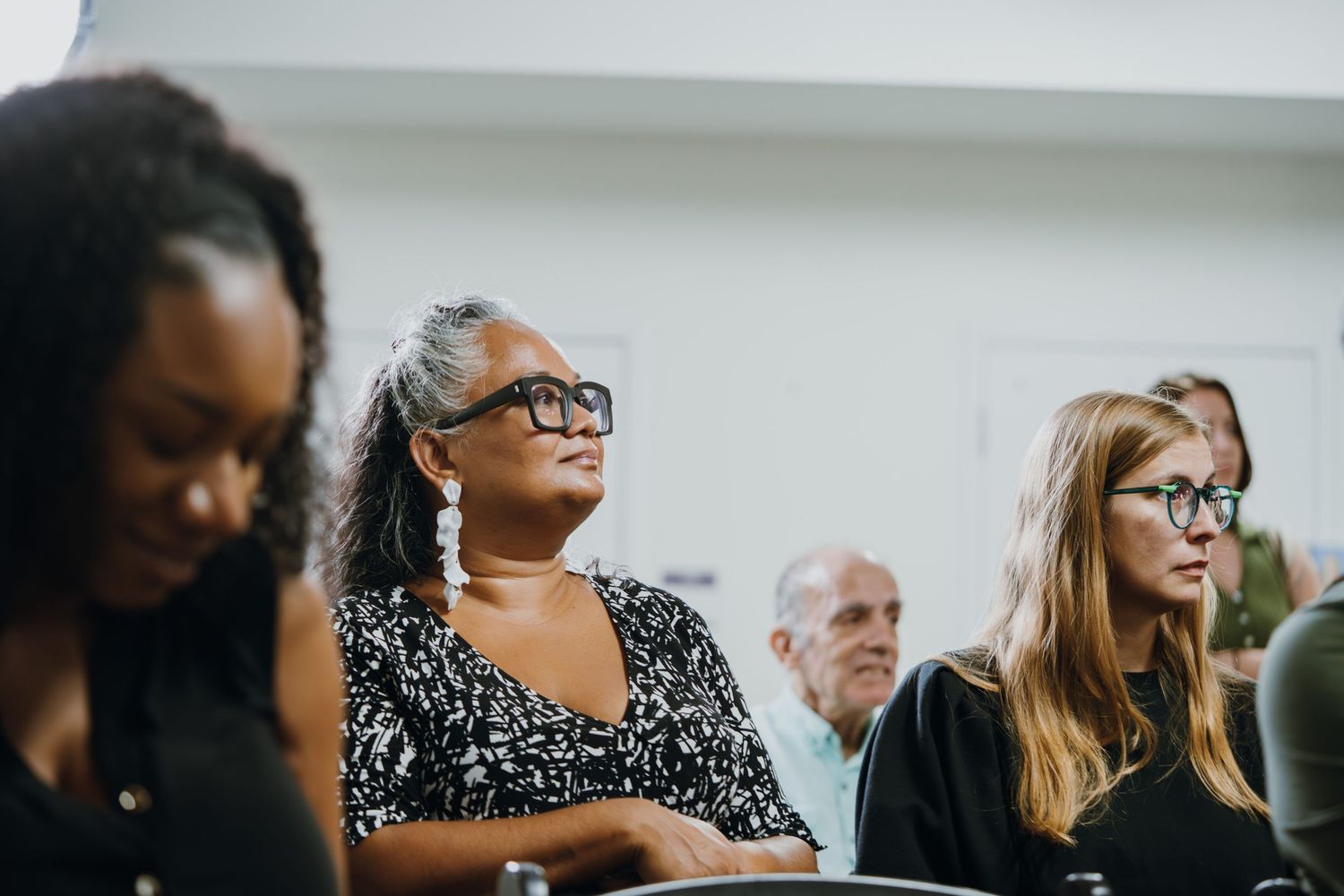 Three diverse audience members attentively listening at an indoor community panel discussion event.