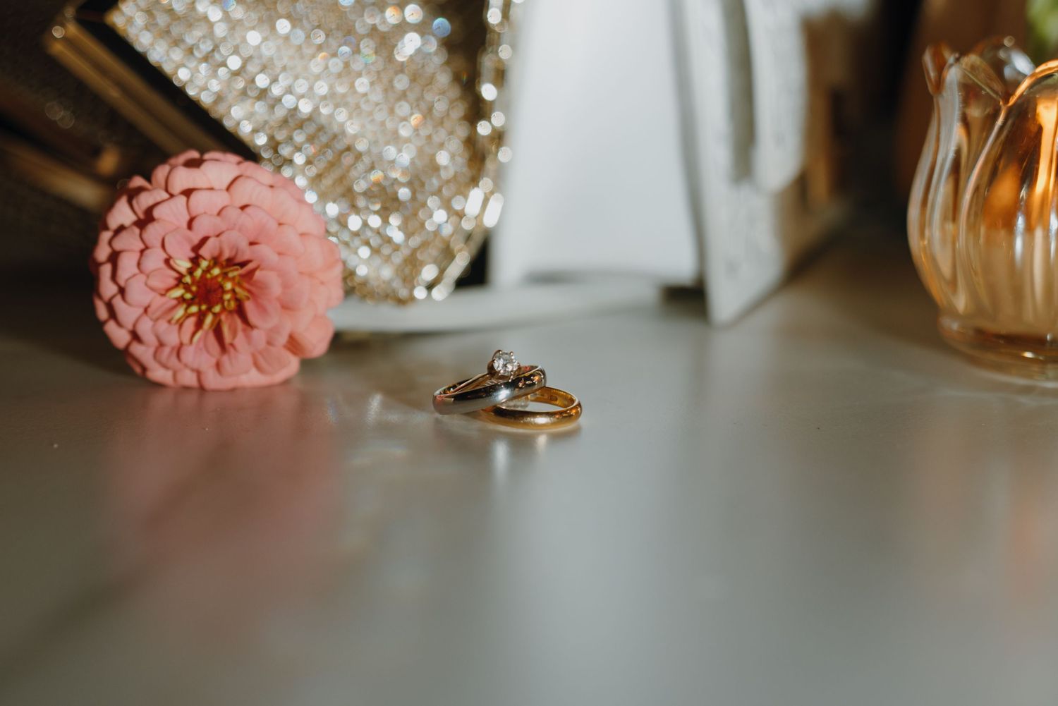 Gold wedding rings with diamond engagement ring displayed beside pink zinnia flower on white surface. New Orleans, Louisiana, Mississippi