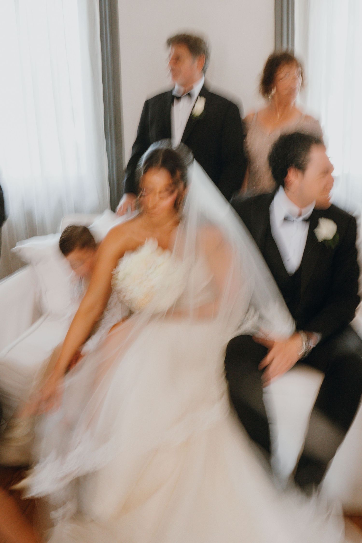 Artistic motion-blur wedding photo of bride in white gown and groom in tuxedo with family in background.