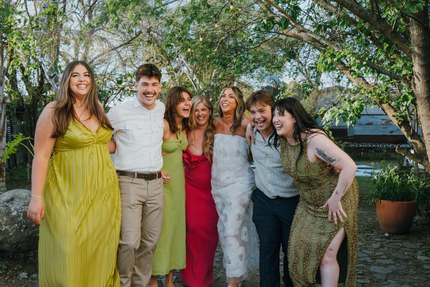 Six happy wedding guests pose together laughing in colorful formal attire at an outdoor garden wedding.