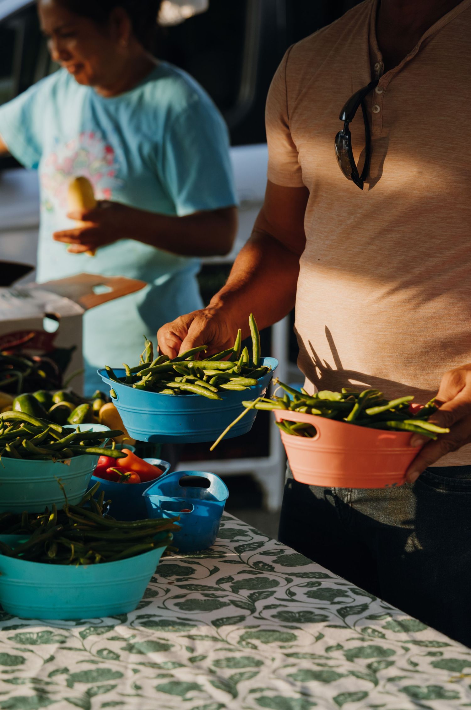 Customers browsing fresh green peppers and vegetables at a market booth.