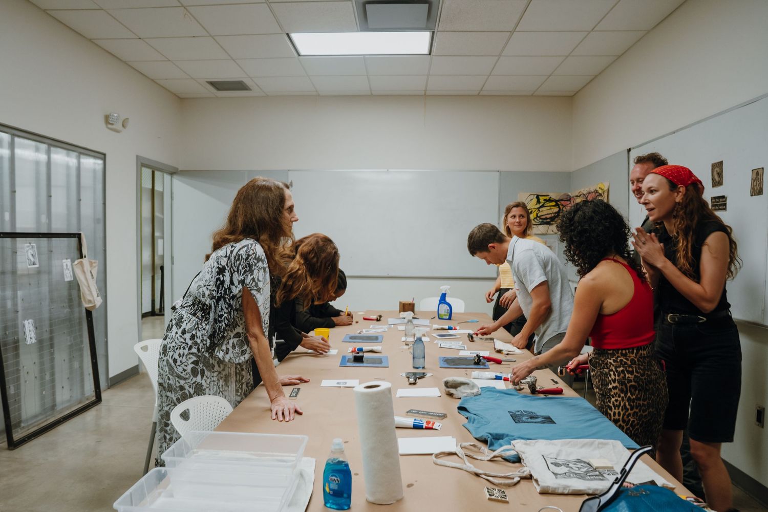 Group of people standing around a long table participating in a hands-on art workshop in a classroom.