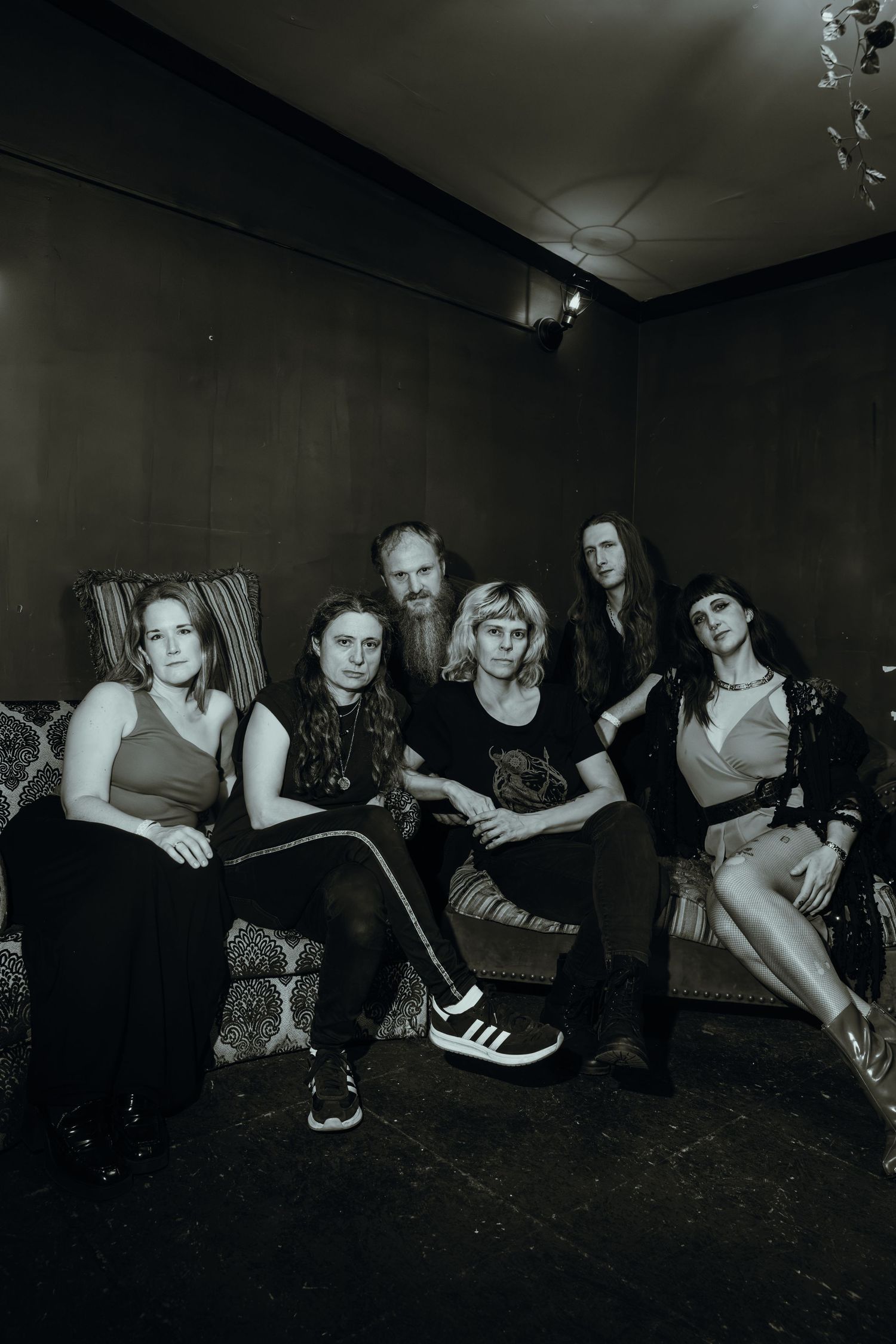 Six theater artists pose together on vintage sofa in moody black and white portrait.