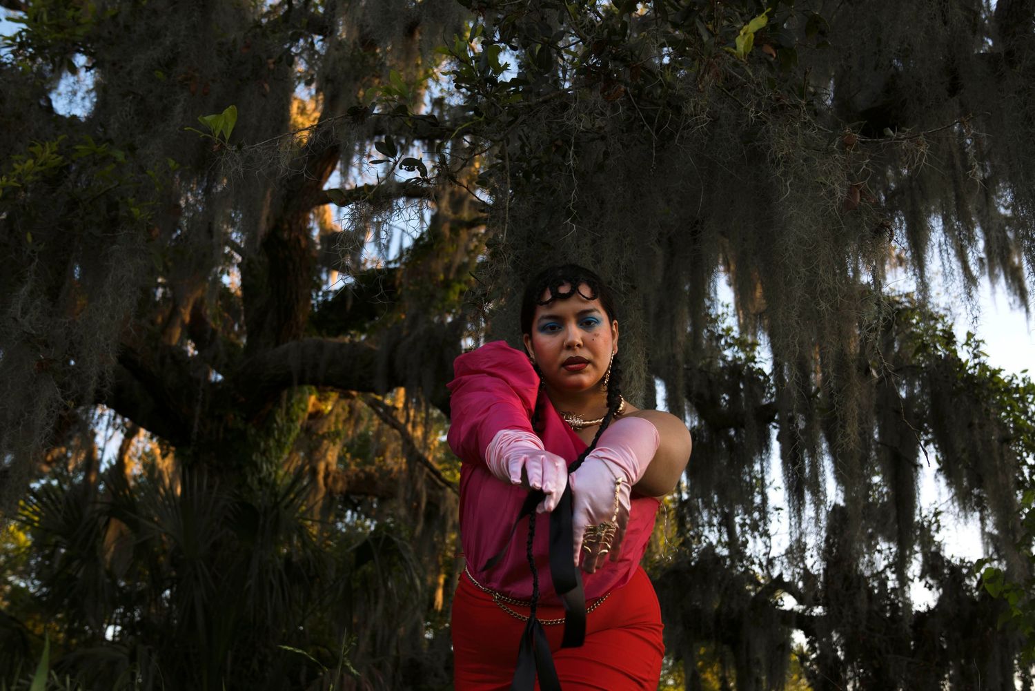 Woman in pink and red outfit reaching toward a tree draped with Spanish moss outdoors.