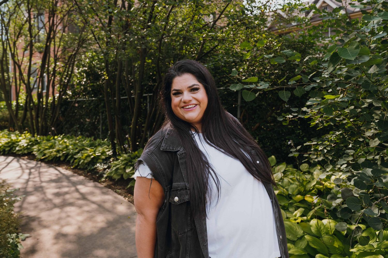 Smiling woman with long dark hair wearing a white tee and dark vest on a garden path.
