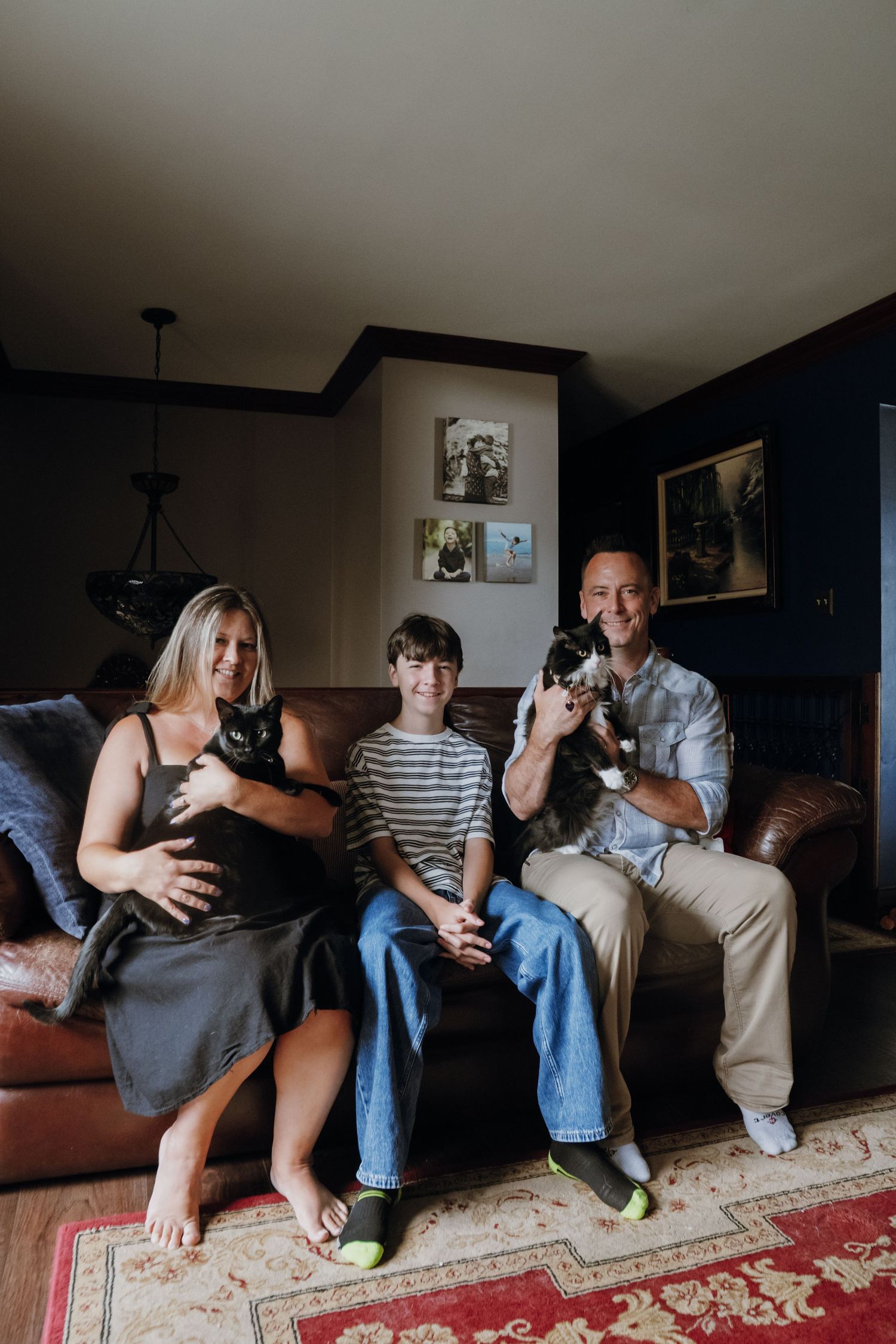 A smiling family of three sits on a leather sofa holding two cats in a cozy dark-walled living room.