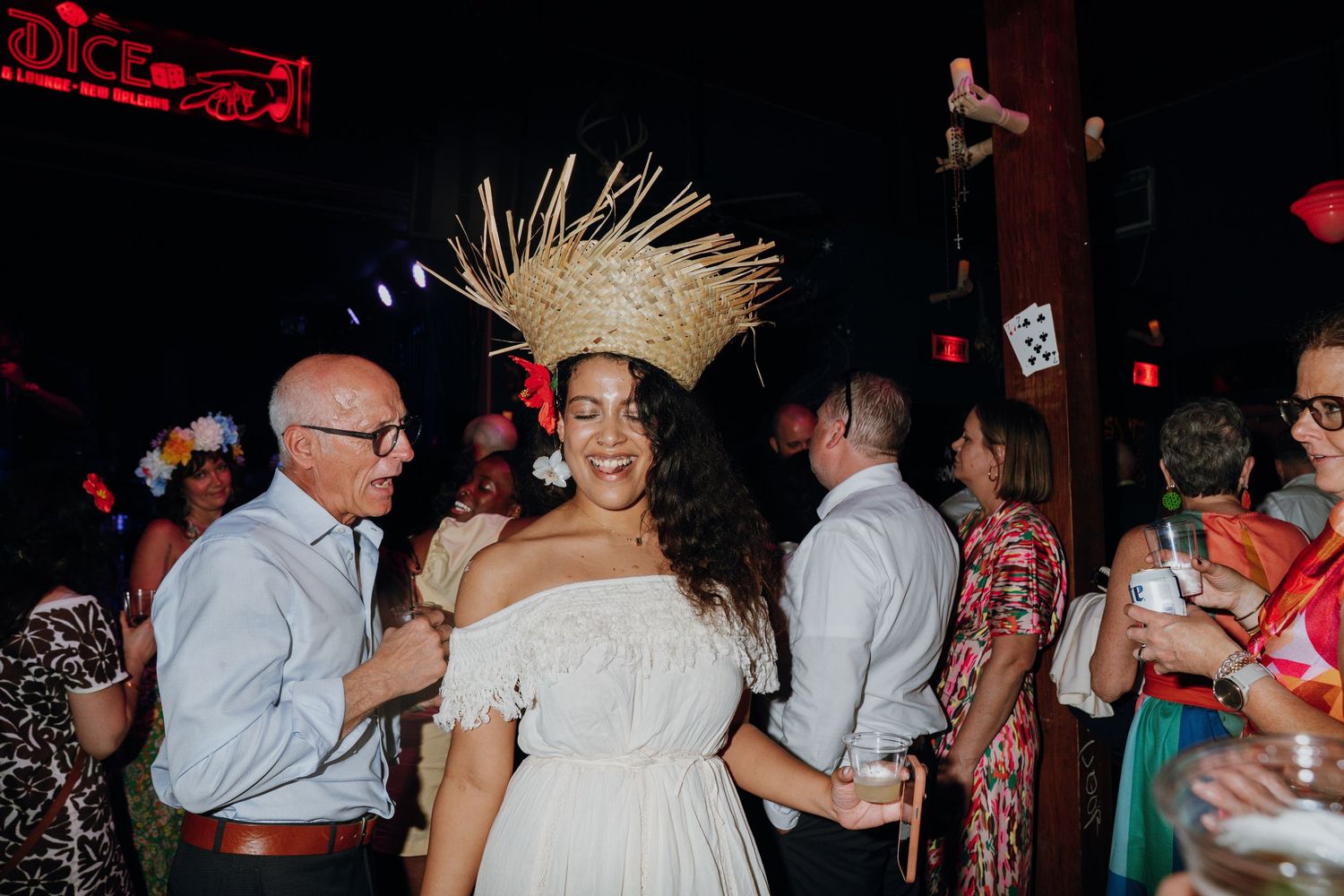 Bride in white off-shoulder dress wearing large straw crown headpiece dancing at festive indoor wedding reception.