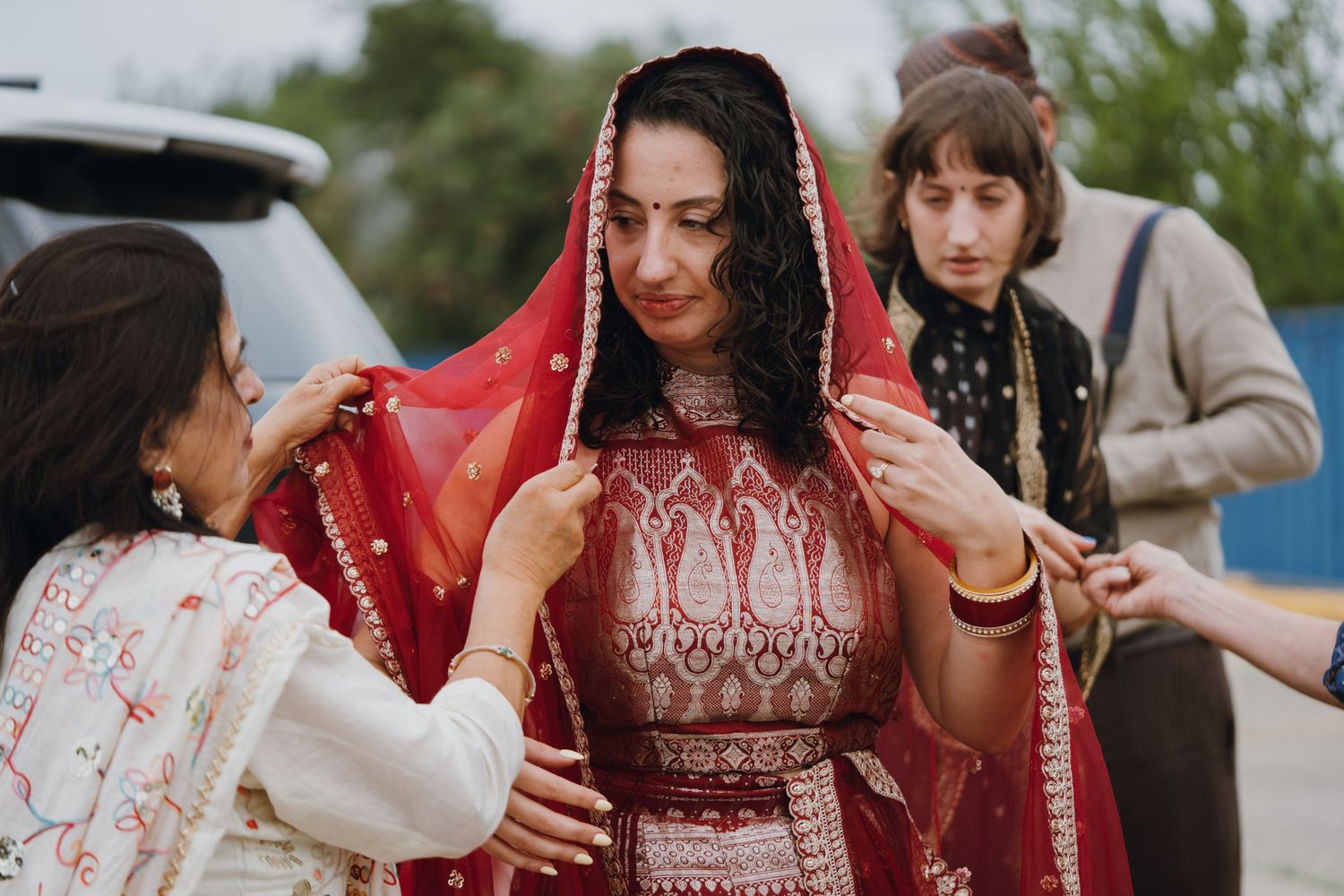 Women help adjust red bridal dupatta veil on South Asian bride in traditional red lehenga.