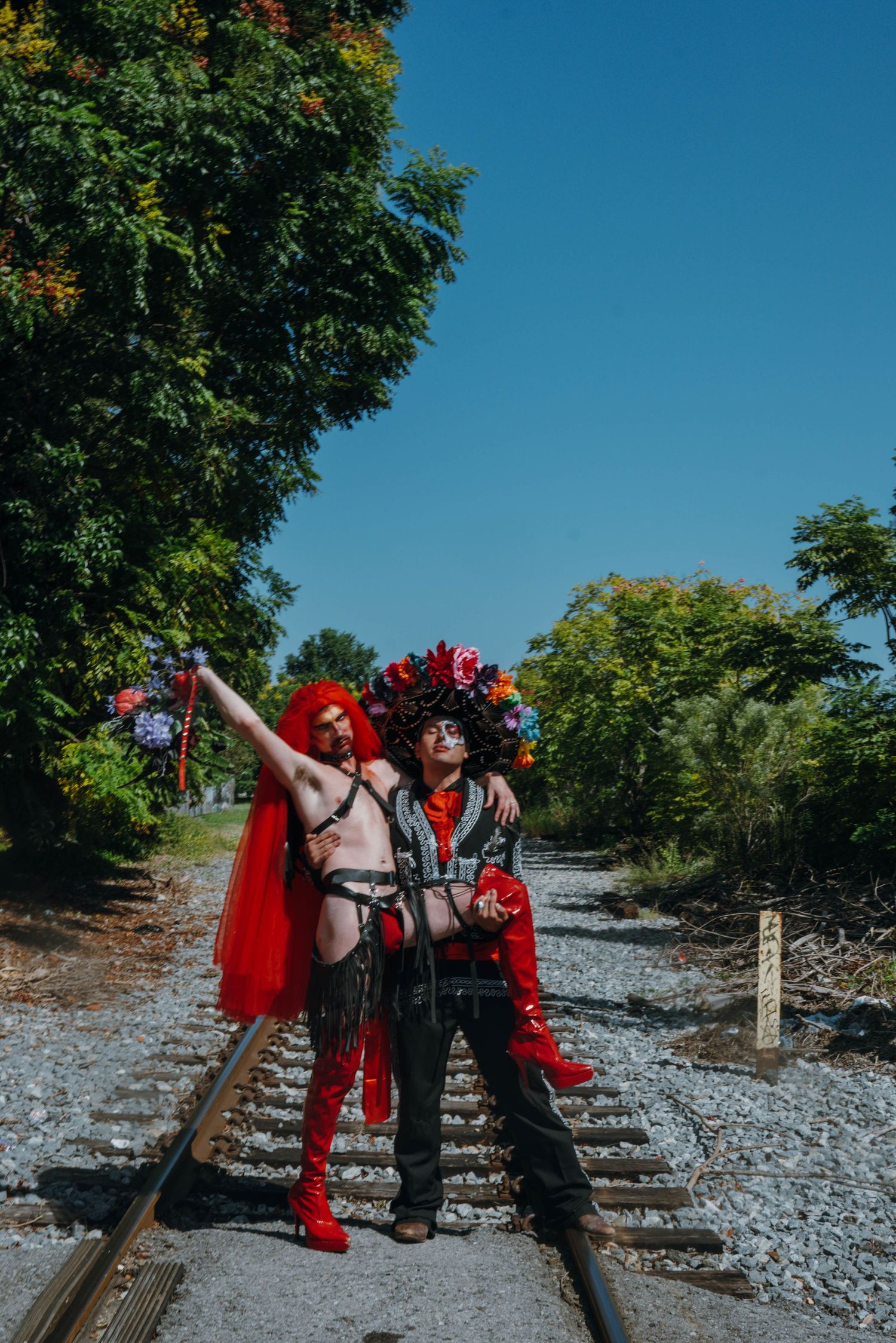 Two costumed performers with floral headpieces posing on railroad tracks on sunny day.