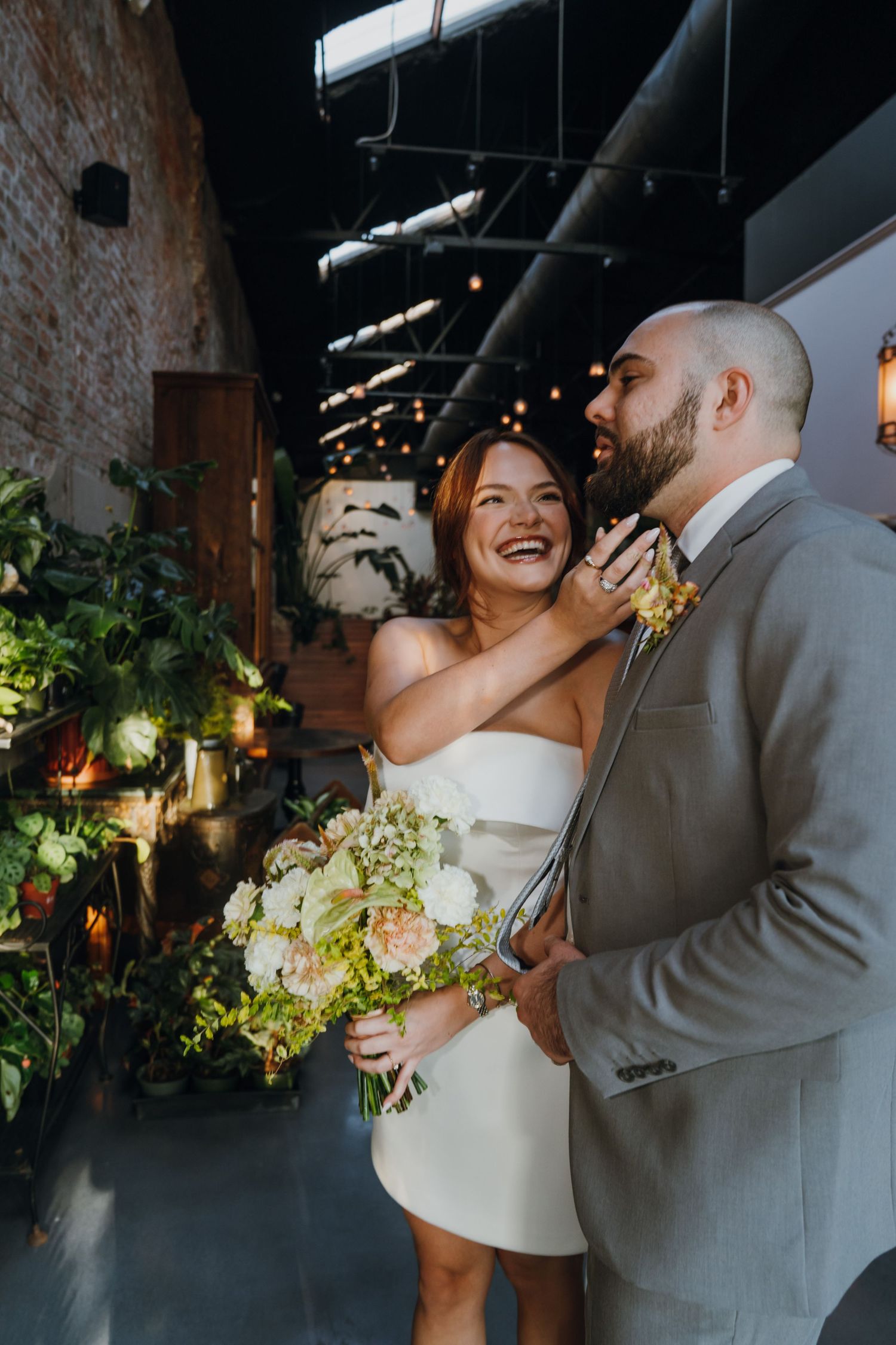 Laughing bride in strapless white mini dress holds bouquet while embracing groom in gray suit indoors. New Orleans, Louisiana, Mississippi