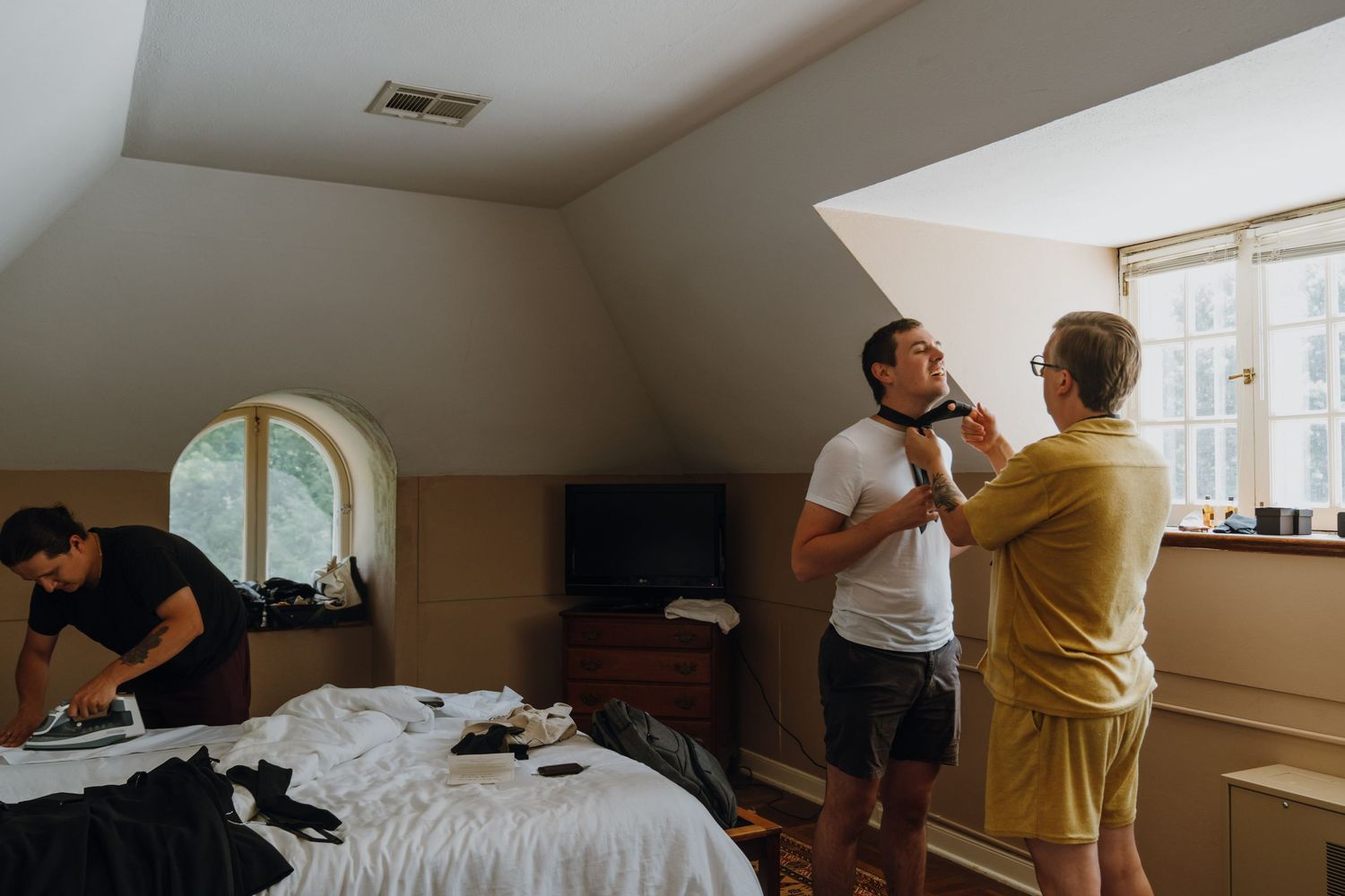 Groomsmen getting ready in attic bedroom, one getting bow tie adjusted near window. New Orleans, Louisiana, Mississippi