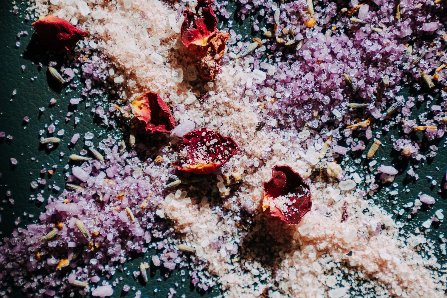 Scattered pink Himalayan salt crystals mixed with dried rose petals and lavender on dark green surface.