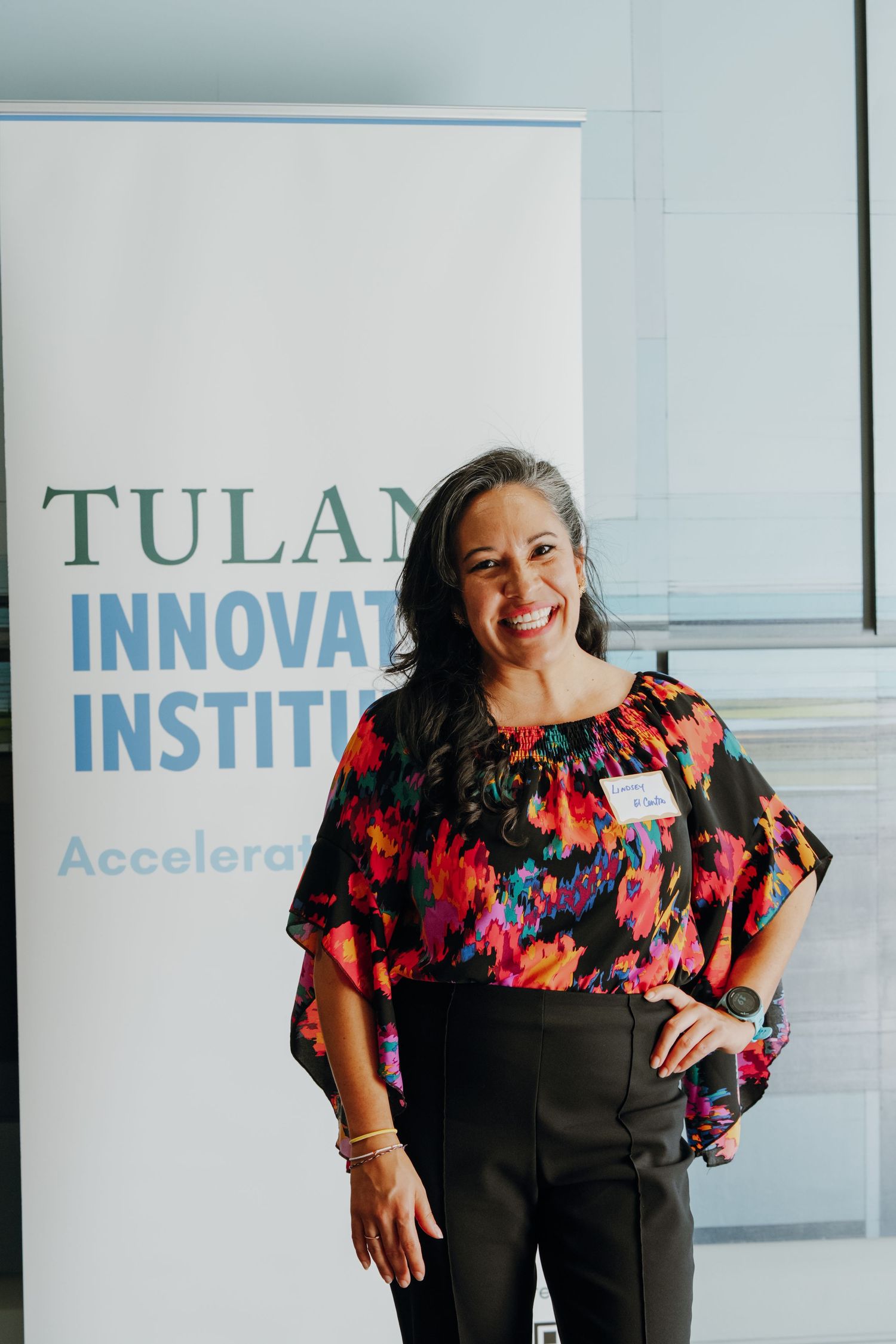 Woman smiling in floral top at Tulane Innovation Institute event, standing before branded banner.