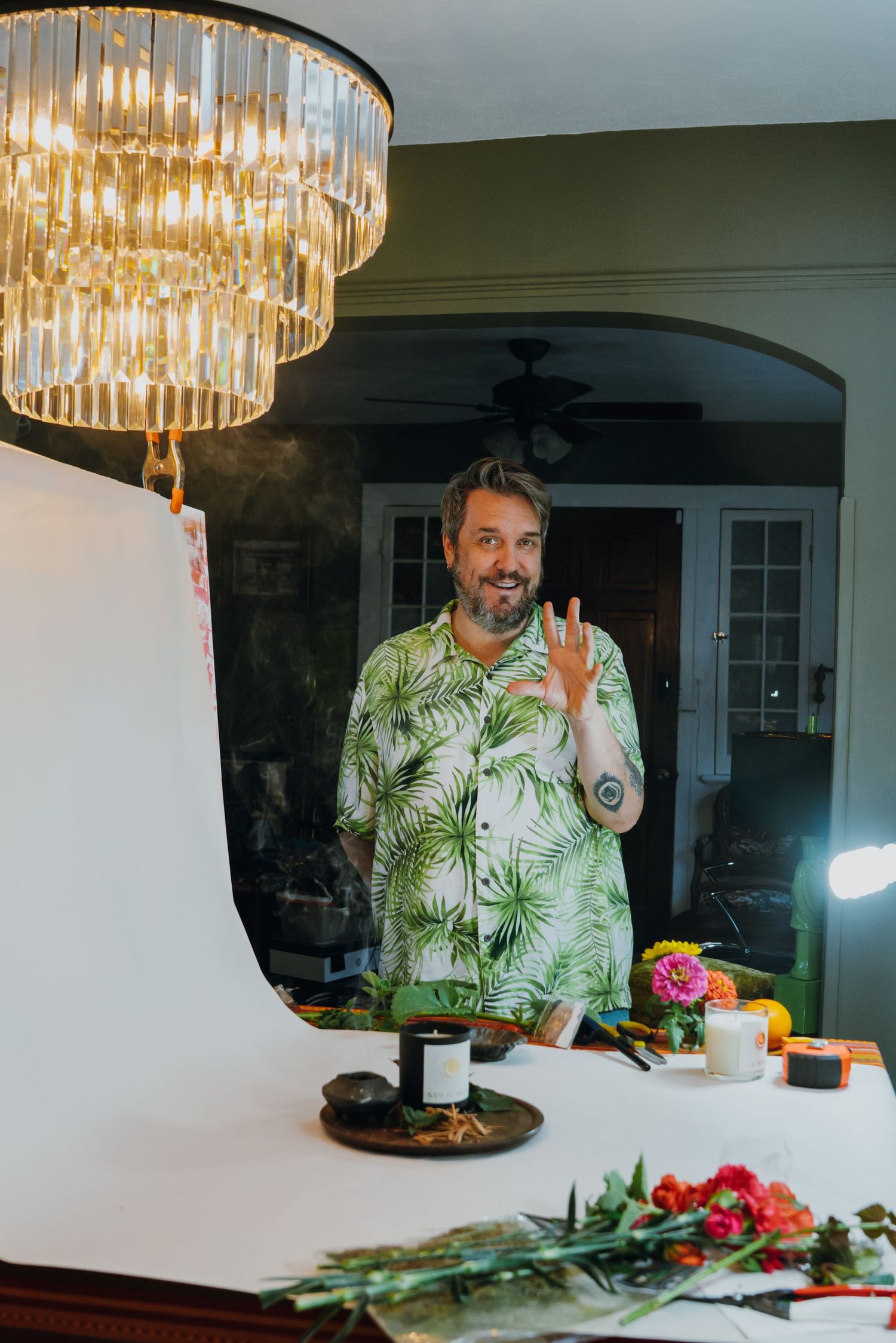 A cheerful man in a tropical shirt waves while standing near a product photography setup.