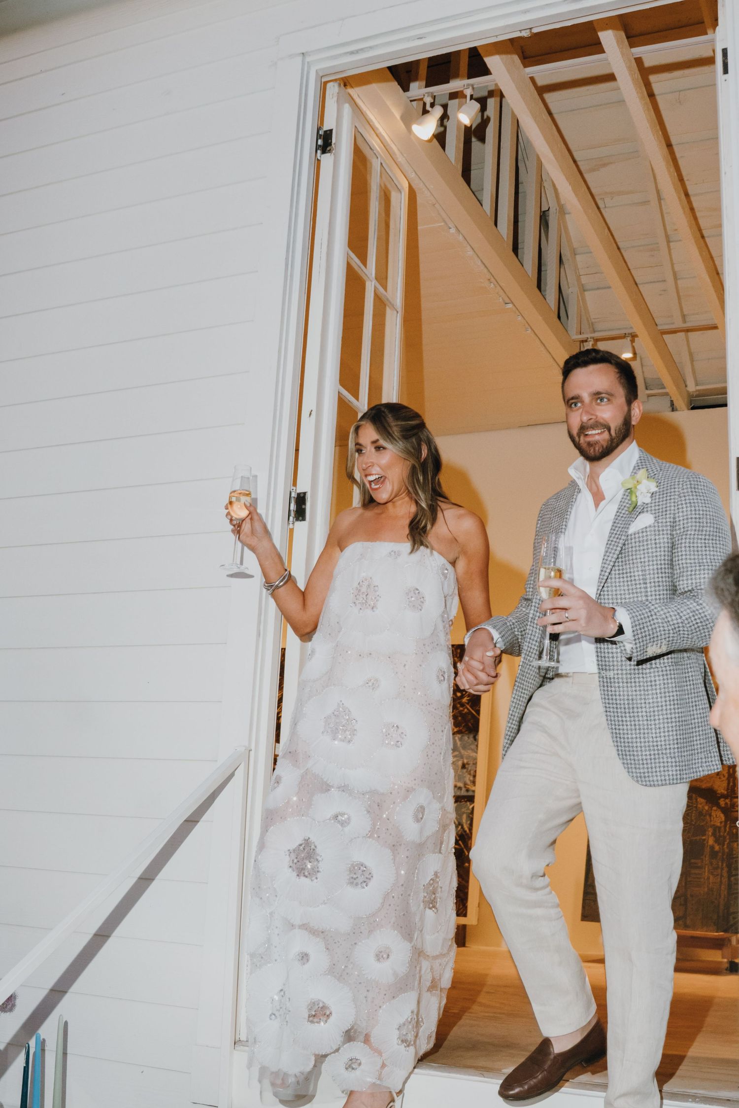 Bride in floral gown and groom in plaid blazer make grand entrance holding champagne glasses.