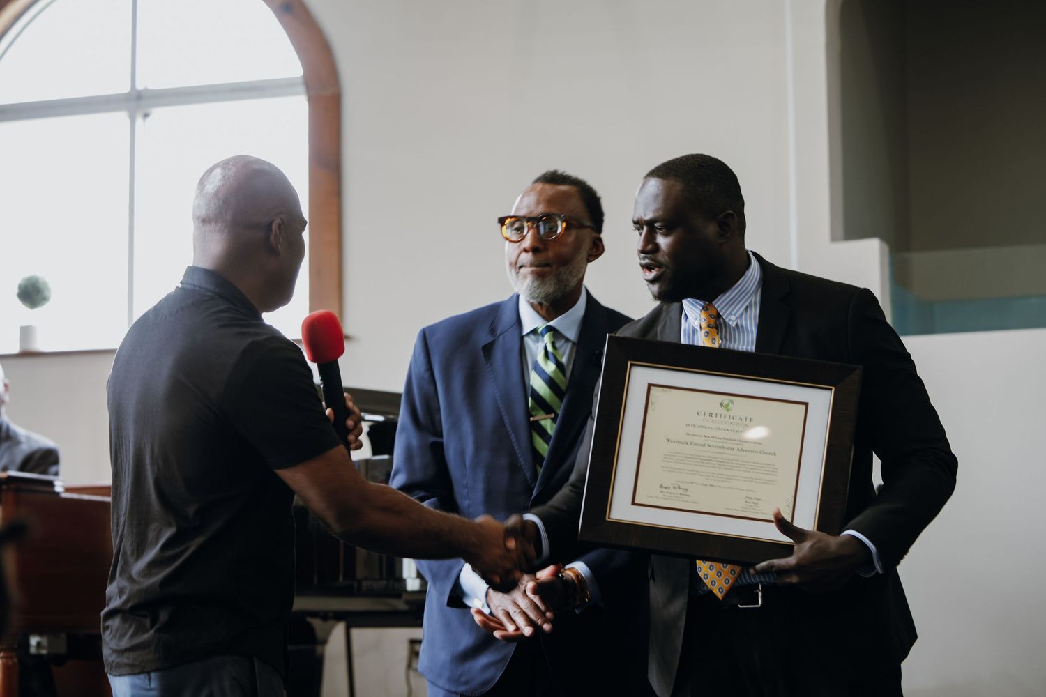 Three men in suits exchanging handshake while holding framed certificate in church setting.