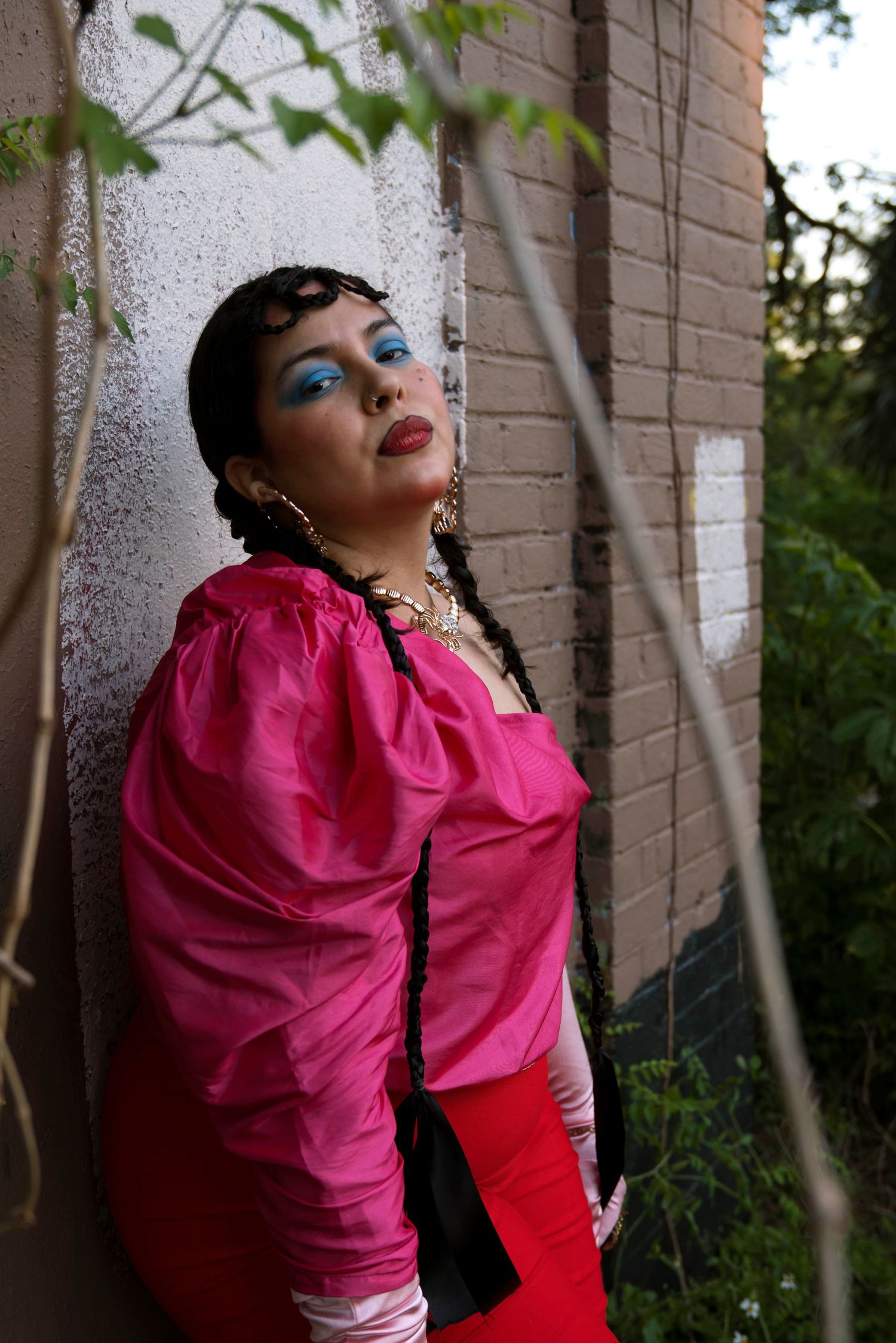 Woman in pink top with blue eyeshadow leaning against a brick wall surrounded by green foliage.