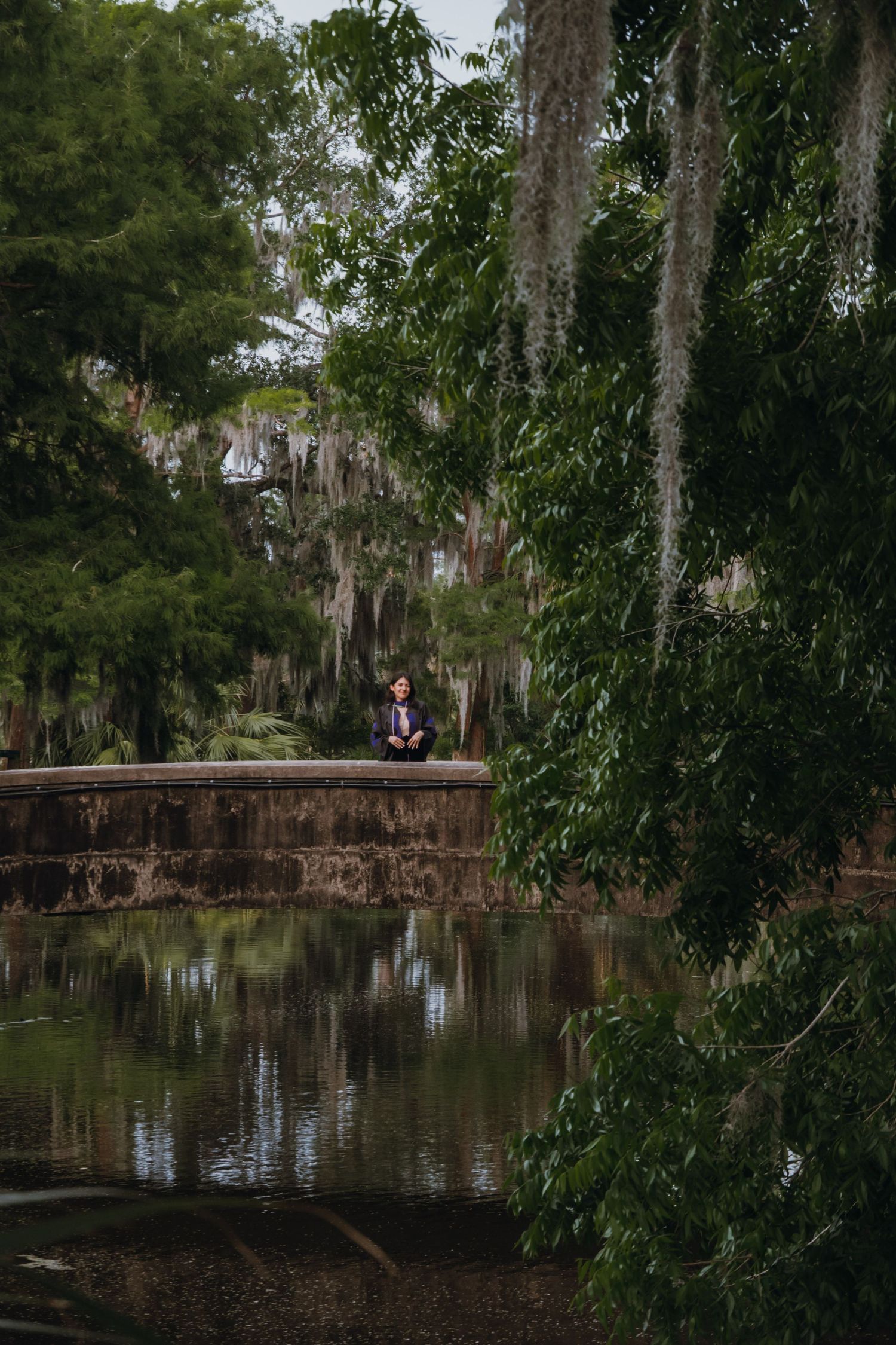 Person sitting on stone wall beside reflective pond surrounded by lush Spanish moss trees.