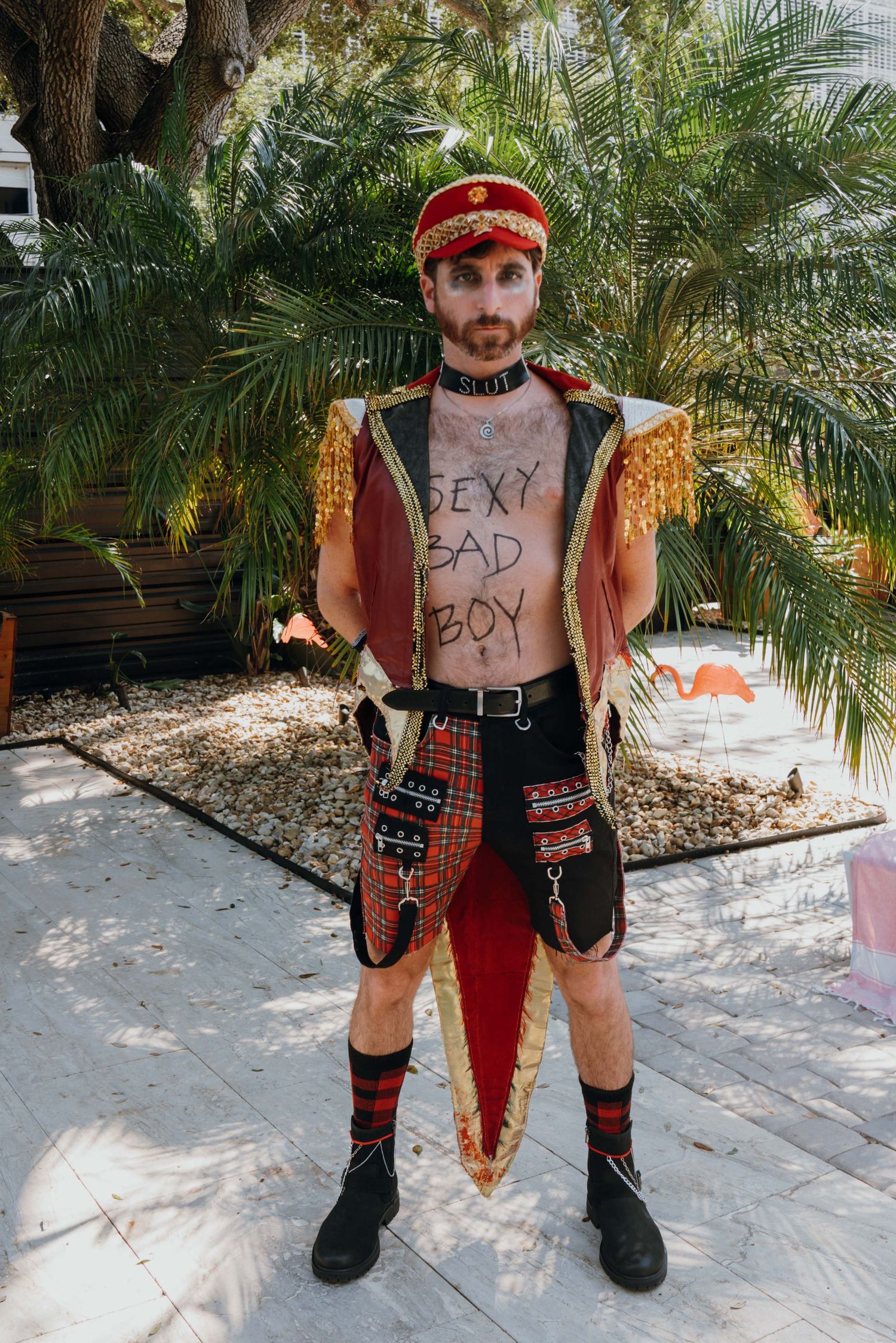 Man in elaborate festive costume with gold epaulettes and red hat with text written on chest outdoors.