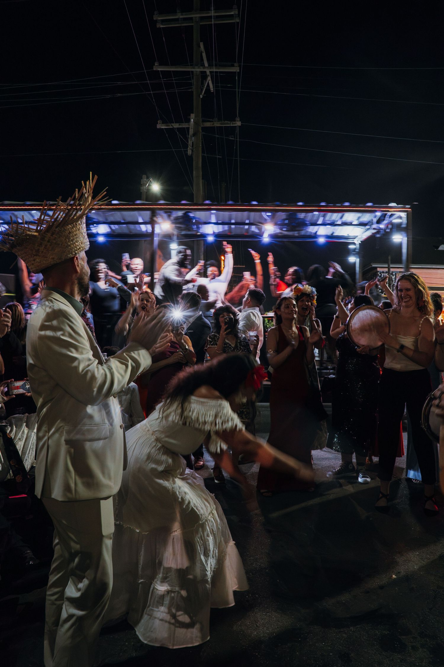 Bride and groom celebrating with large crowd of cheering guests at vibrant outdoor nighttime wedding party.
