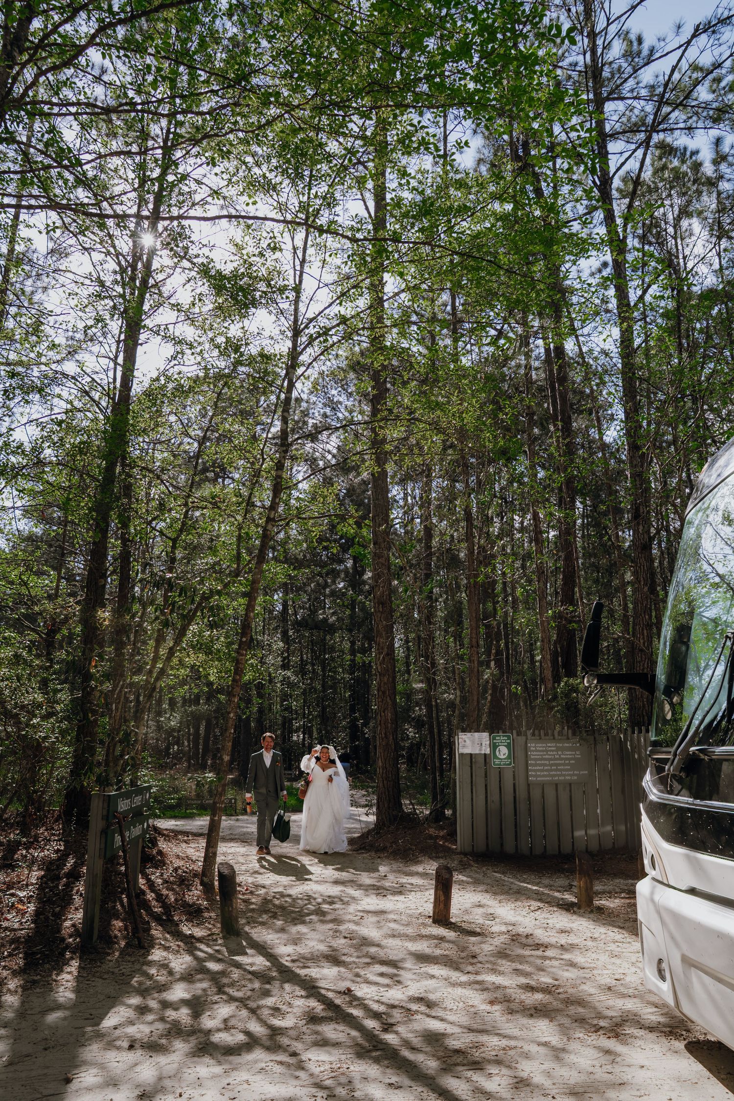 Bride and groom walk along a forest path near a white bus on their wedding day.