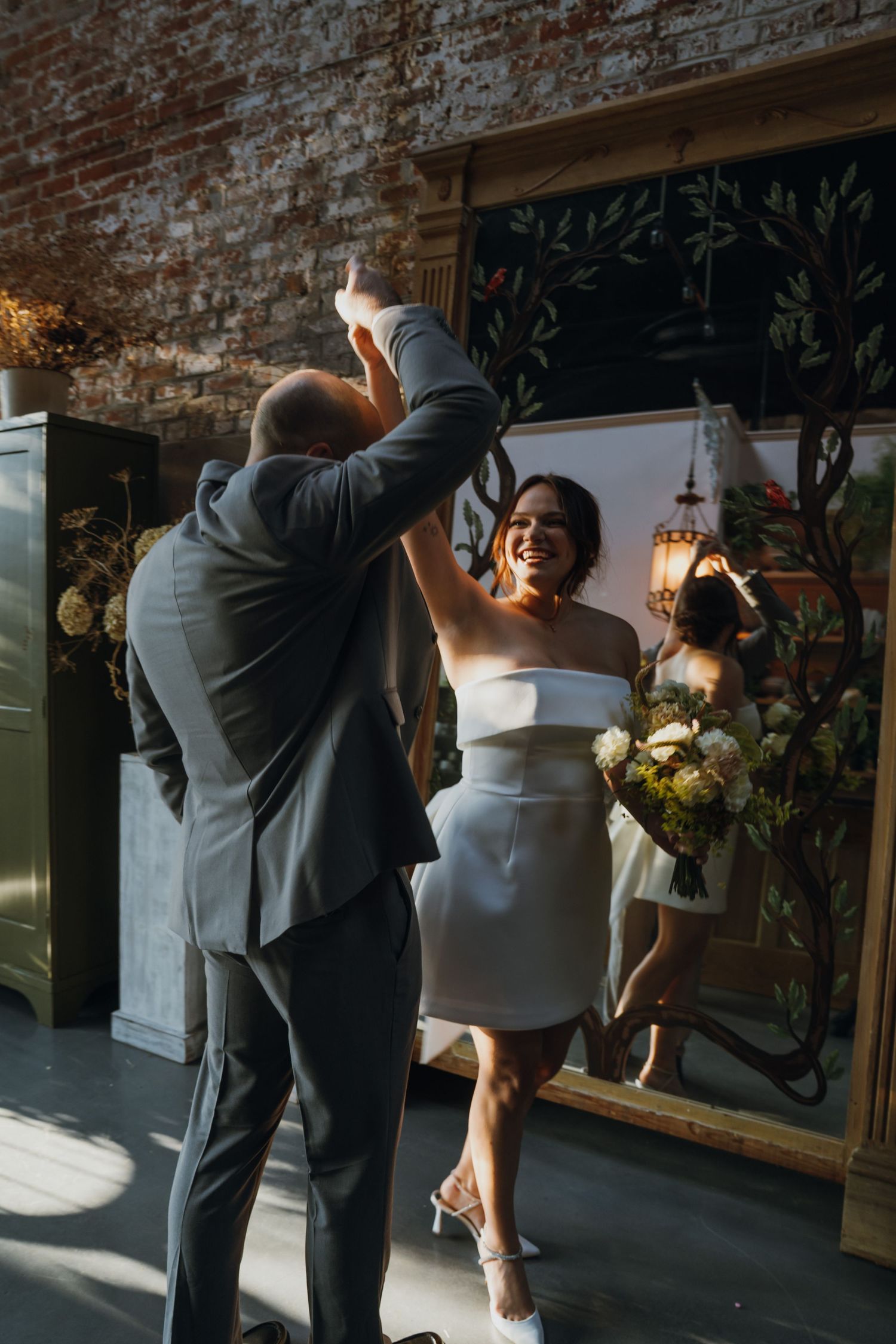 Joyful bride in white mini dress spins with guest near decorative mirror at indoor wedding celebration. New Orleans, Louisiana, Mississippi
