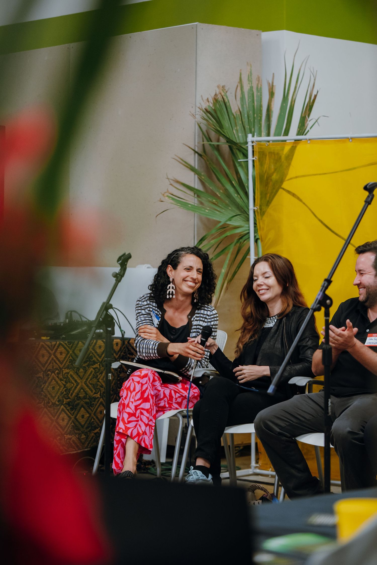 Three panelists laughing during outdoor event, woman in colorful pink floral pants on stage.