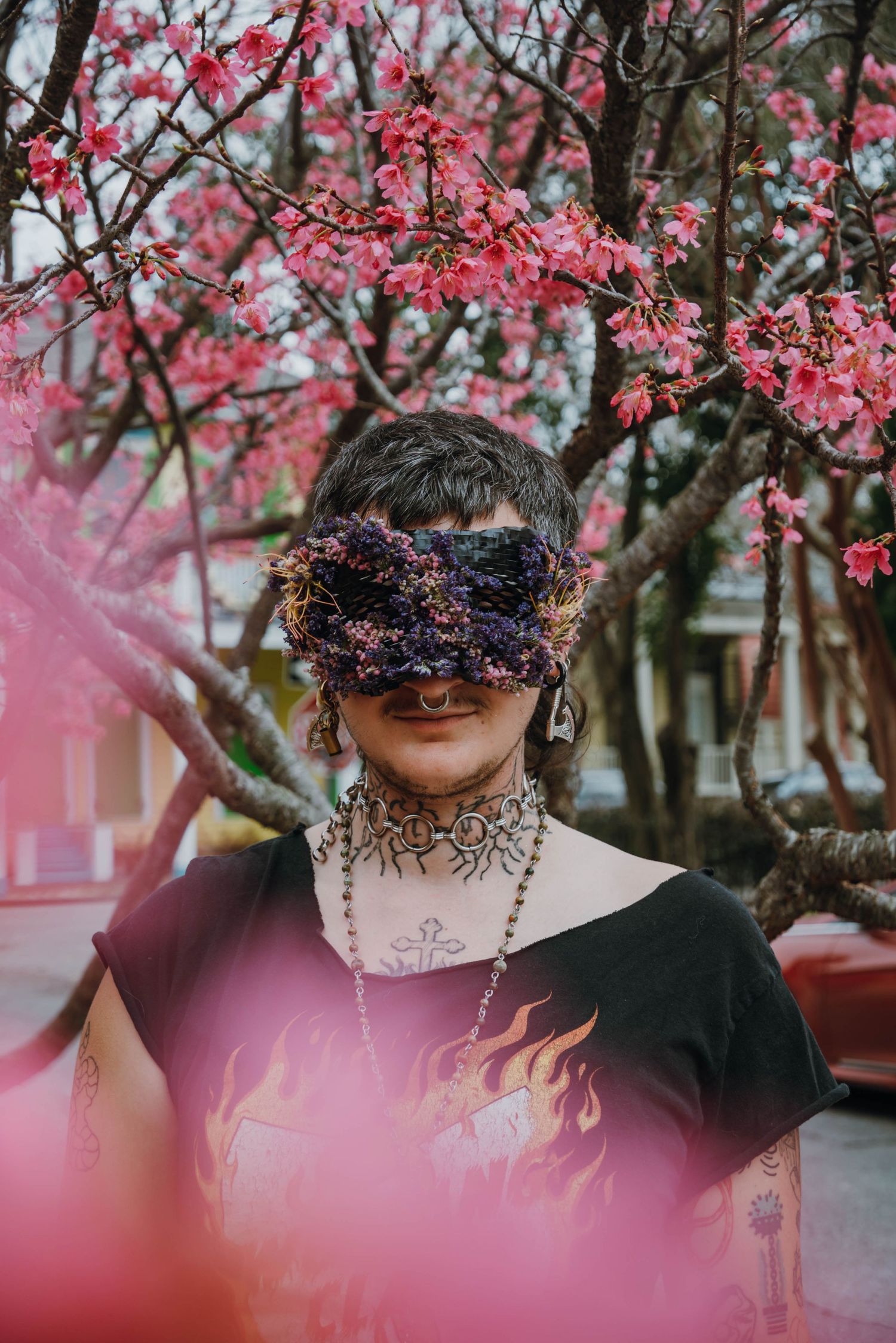 Tattooed person wearing a floral mask standing under blooming pink cherry blossom trees outdoors.