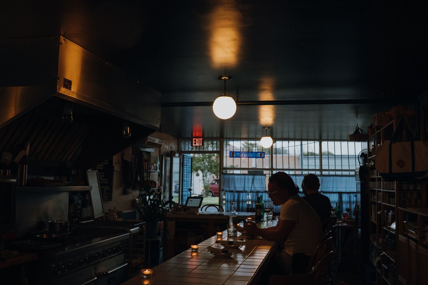 Dimly lit restaurant interior with bar seating, pendant lights, and a kitchen visible in the background.