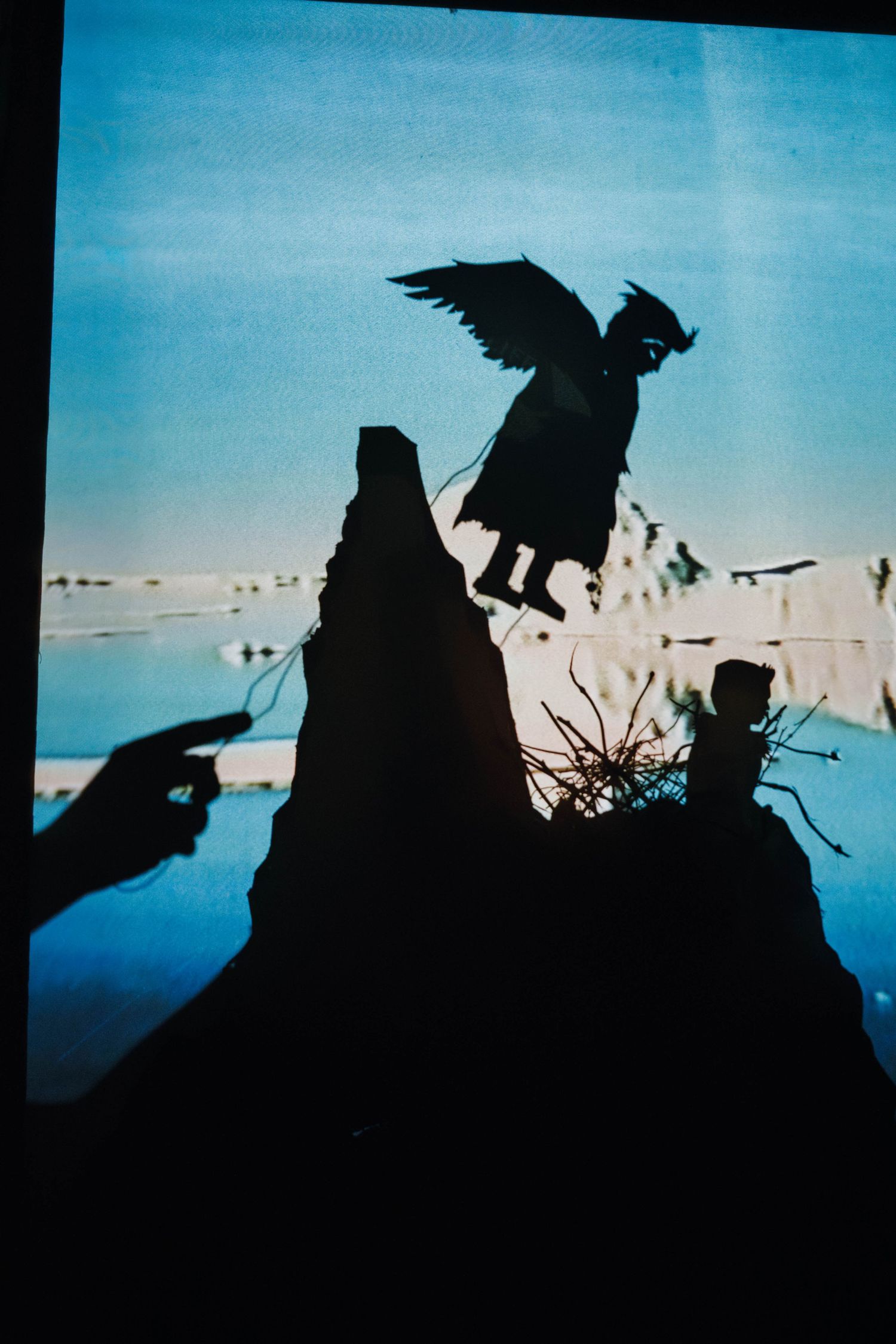 Shadow puppet silhouette of winged figure soaring over rocky landscape projected on blue screen.