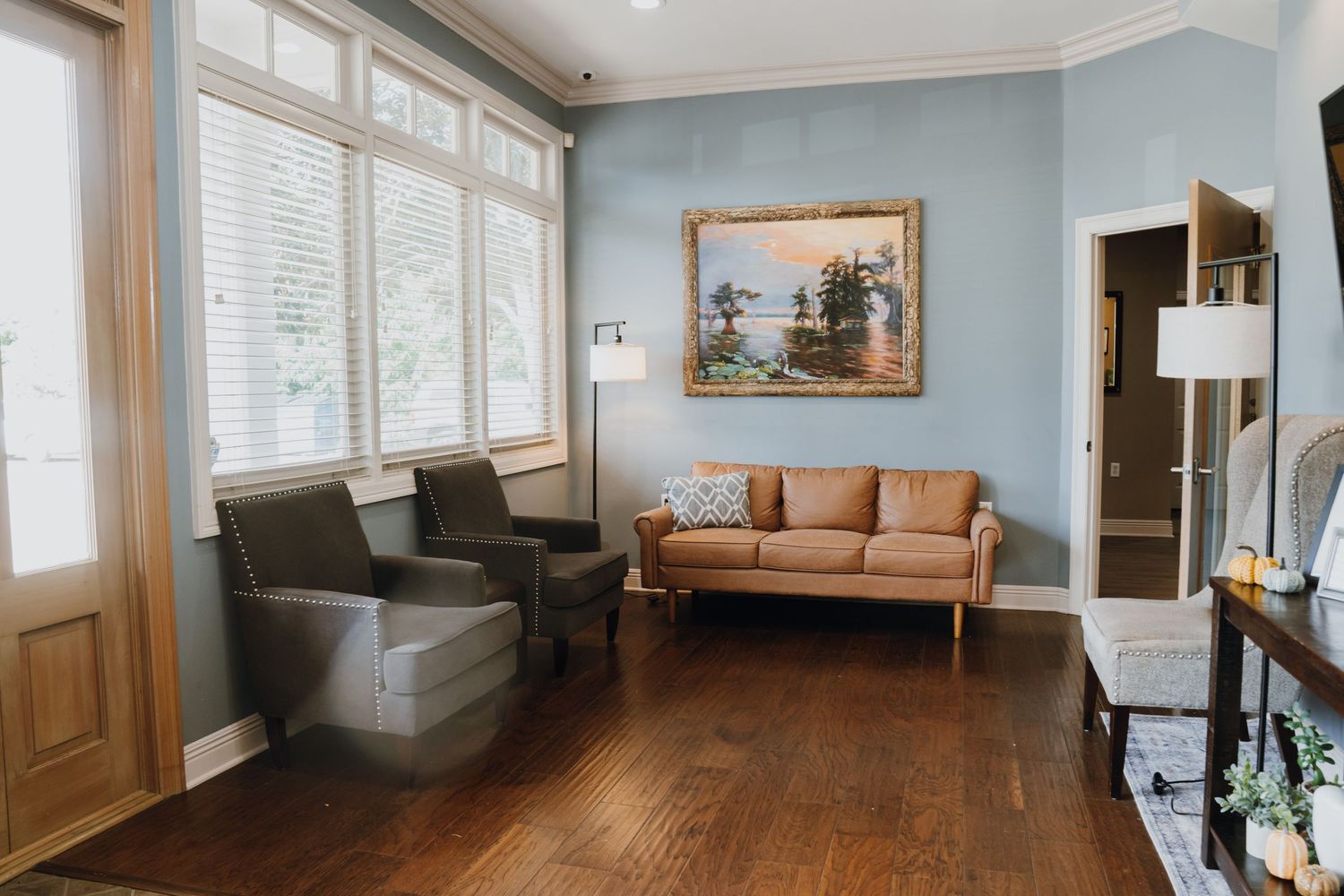 Cozy living room with hardwood floors, tan sofa, gray chairs, and landscape painting on blue wall.