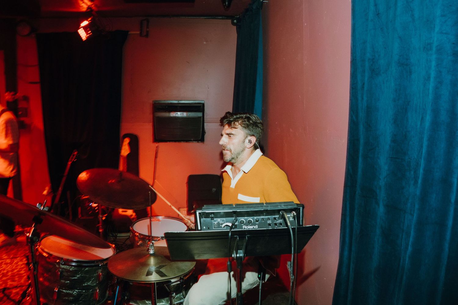 A drummer in an orange sweater plays drums under colorful stage lighting at a live event.