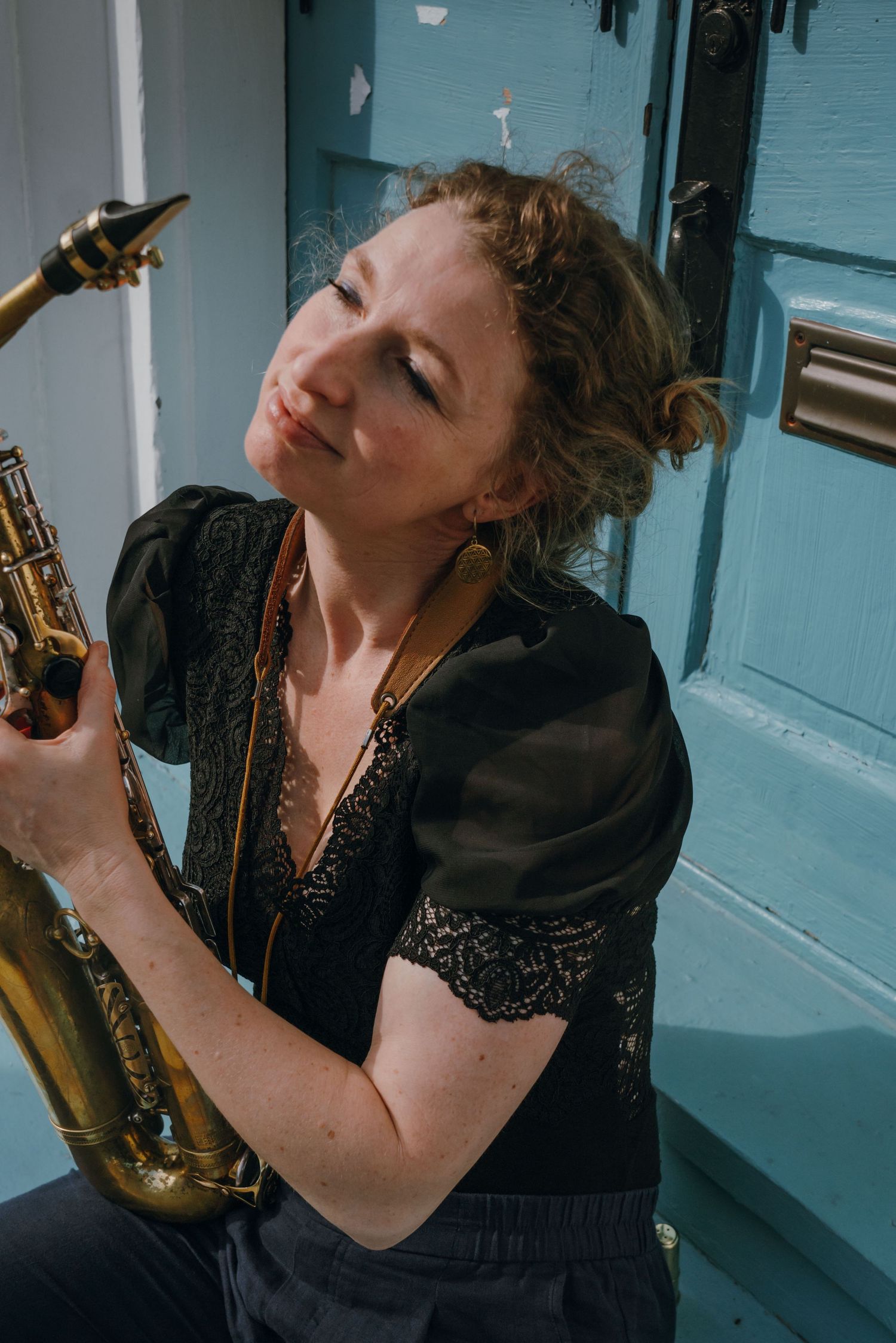 Female musician with curly hair holding brass saxophone while sitting against a light blue door.