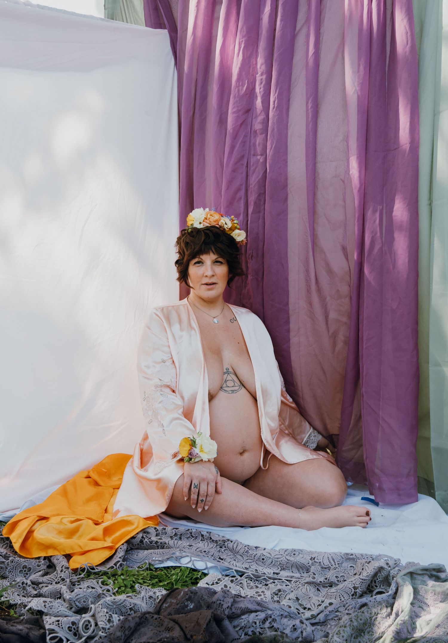 Pregnant woman in floral crown and pink robe sitting on ground with orange and gray fabrics against purple drapes.