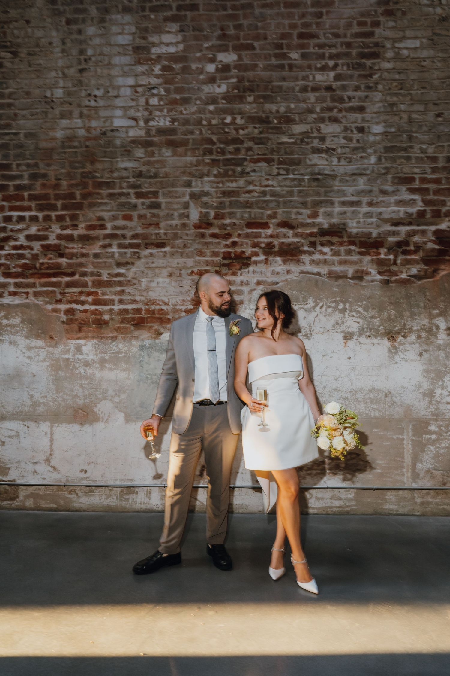 Bride and groom pose together holding champagne against rustic exposed brick wall in golden sunlight. New Orleans, Louisiana, Mississippi