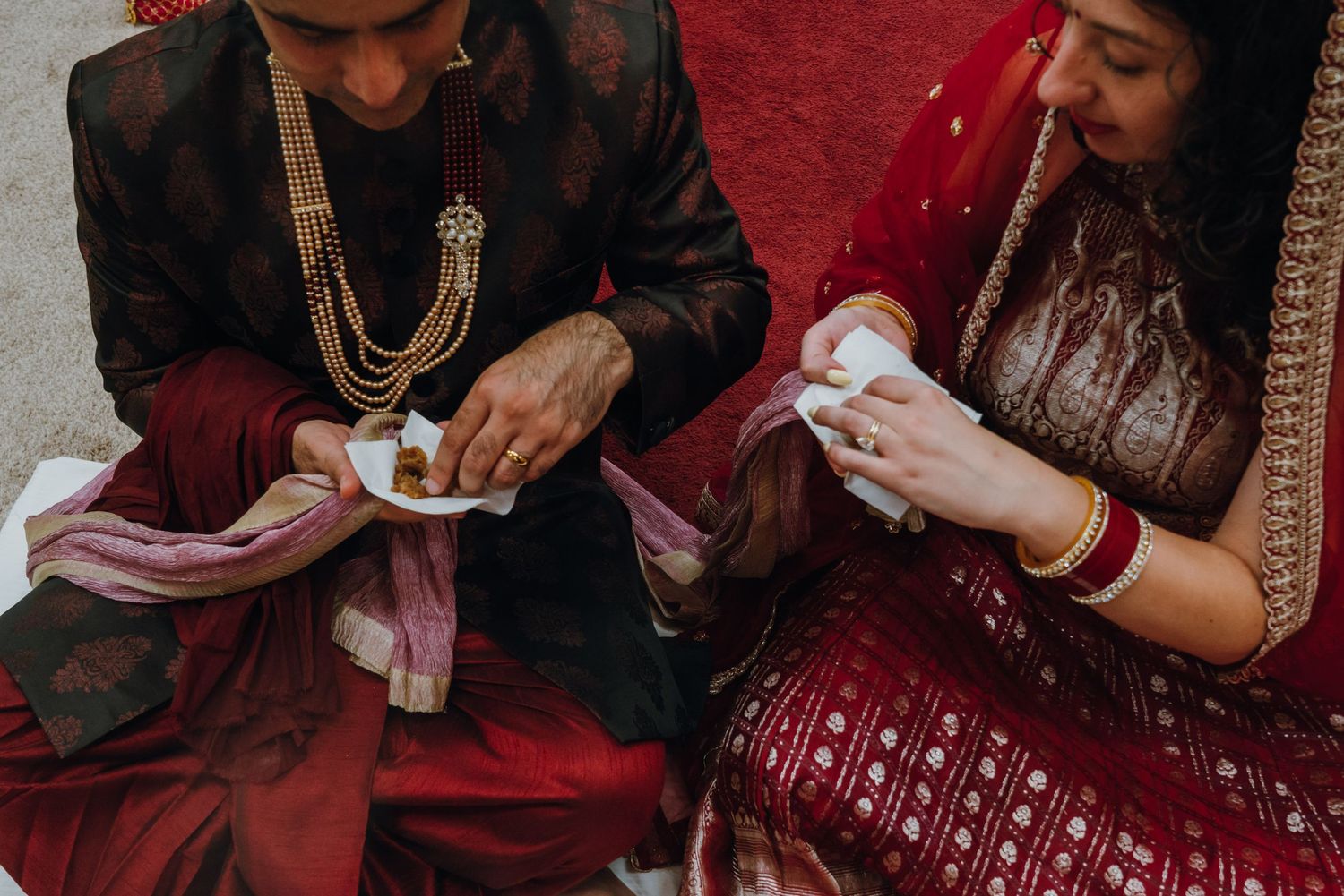 Indian bride and groom in traditional red and dark attire sharing sweets during wedding ceremony ritual.